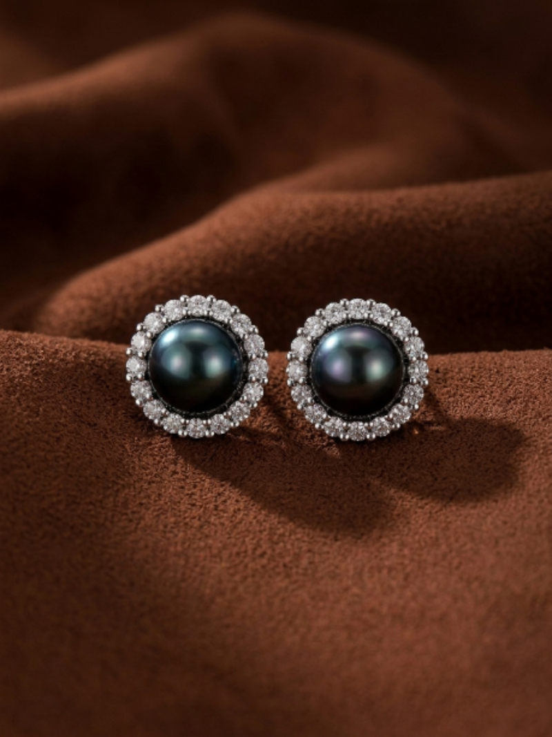 Close-up of two Shadowlight Tahitian Pearl halo stud earrings featuring a 10mm round black pearl surrounded by a halo of sparkling accent stones, set against a textured brown background.