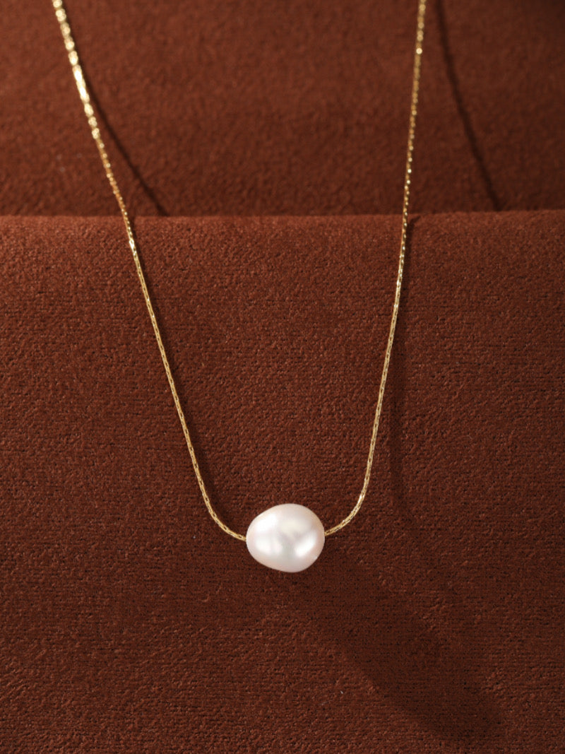 Solace Freshwater Pearl Small Pendant Necklace featuring a lustrous 6 mm white seed pearl on a delicate gold-toned chain against a brown textured background.