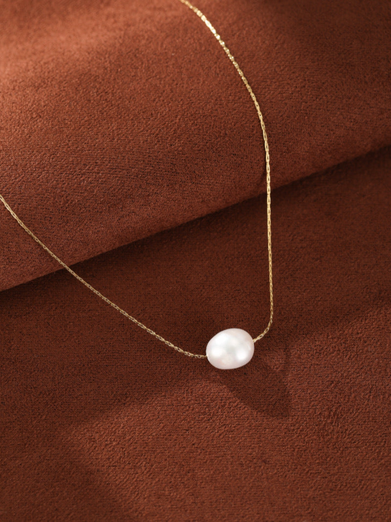 A minimalist gold-toned chain necklace featuring a single, lustrous 6mm white freshwater seed pearl pendant on a textured brown background.
