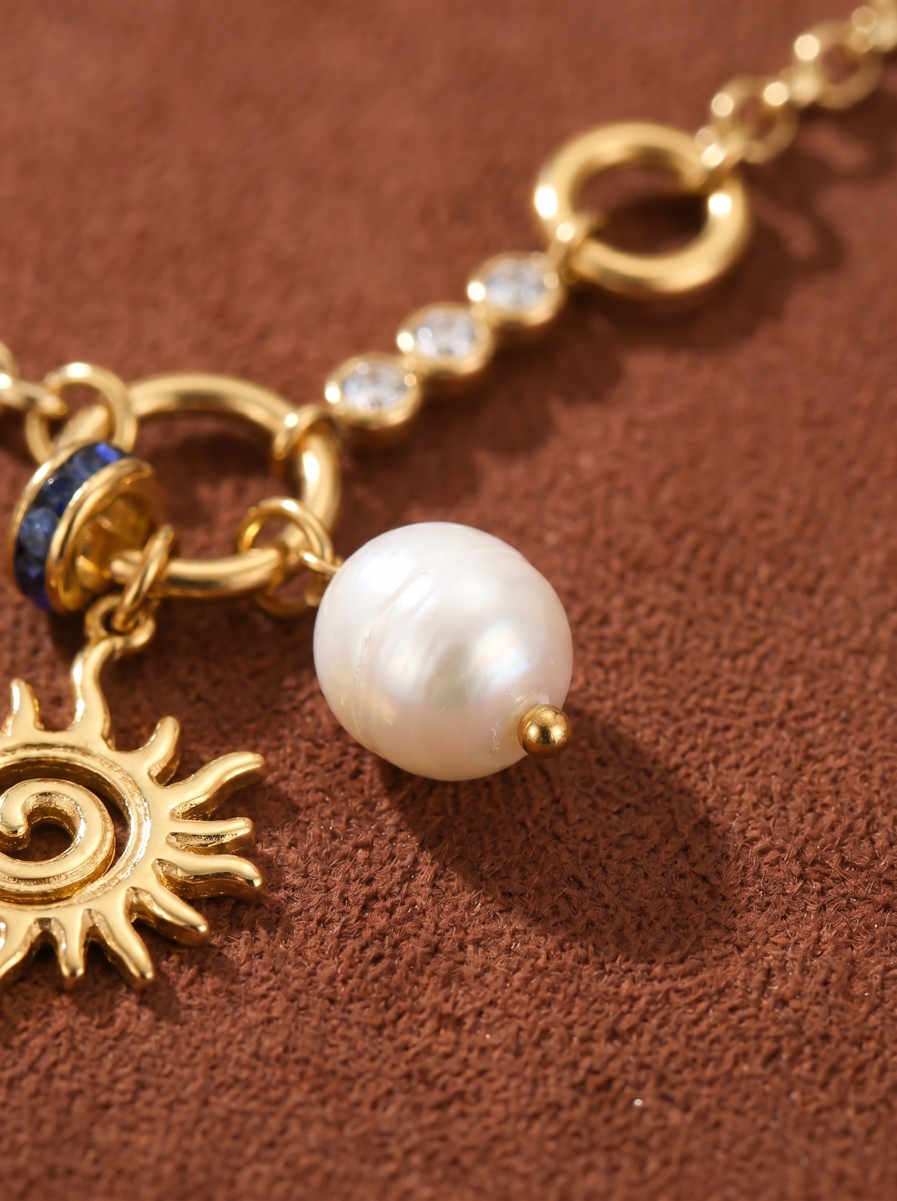 Close-up of a gold-tone chain necklace featuring a sun pendant, a lustrous white freshwater pearl, and a blue accent bead, with clear stones along the chain. @pearl