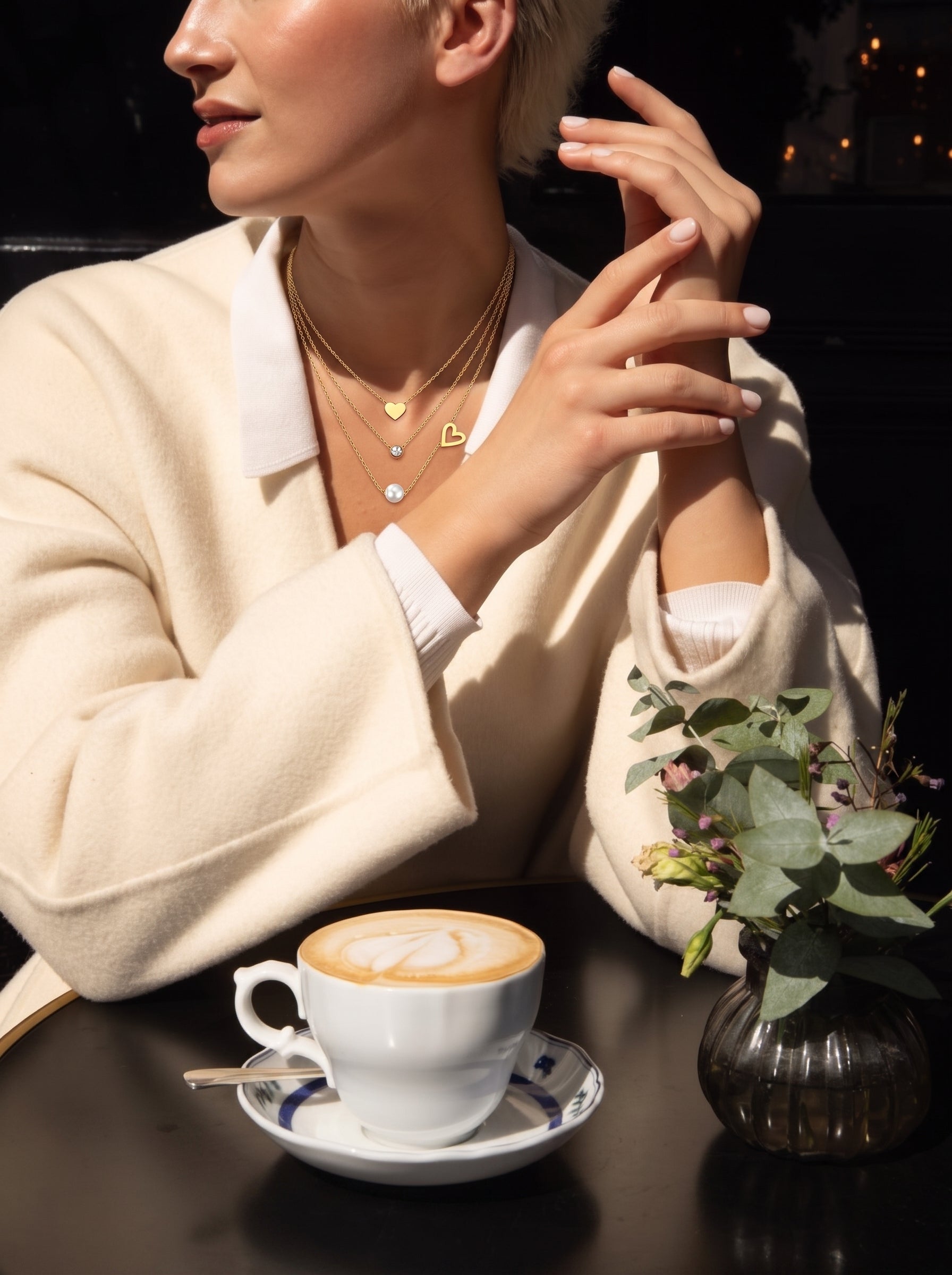 Une femme portant un manteau crème et un collier superposé présentant une breloque cœur, une pierre accent transparente, et une perle blanche d'eau douce, savoure un café dans un café. @lifestyle