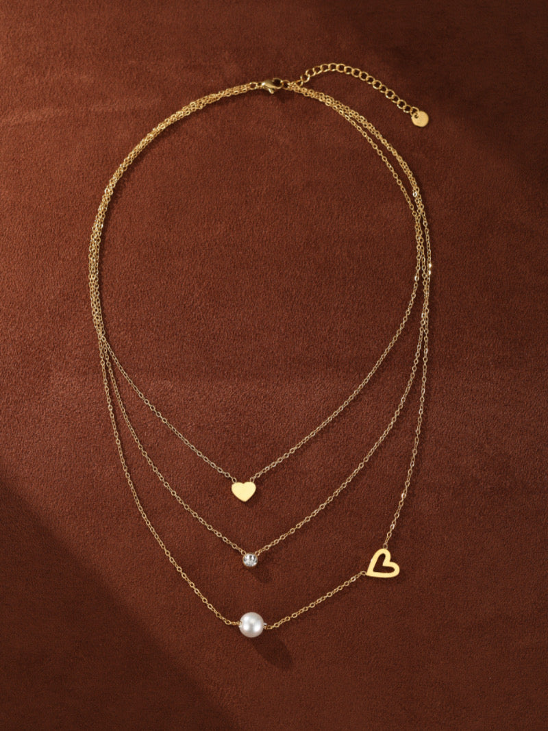 Collier superposé doré avec trois chaînes, présentant une petite breloque cœur, une pierre accent scintillante, et une lustrée perle blanche d'eau douce, ainsi qu'une breloque cœur ouvert sur la chaîne la plus longue.