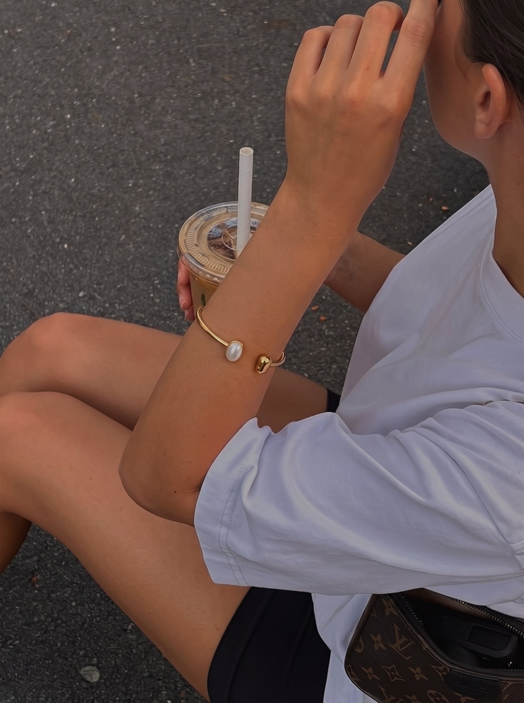 A woman wearing the Span Freshwater Pearl Cuff Bracelet, holding a coffee cup, outdoors. The bracelet features a gold cuff with a baroque pearl and a gold sphere. @lifestyle