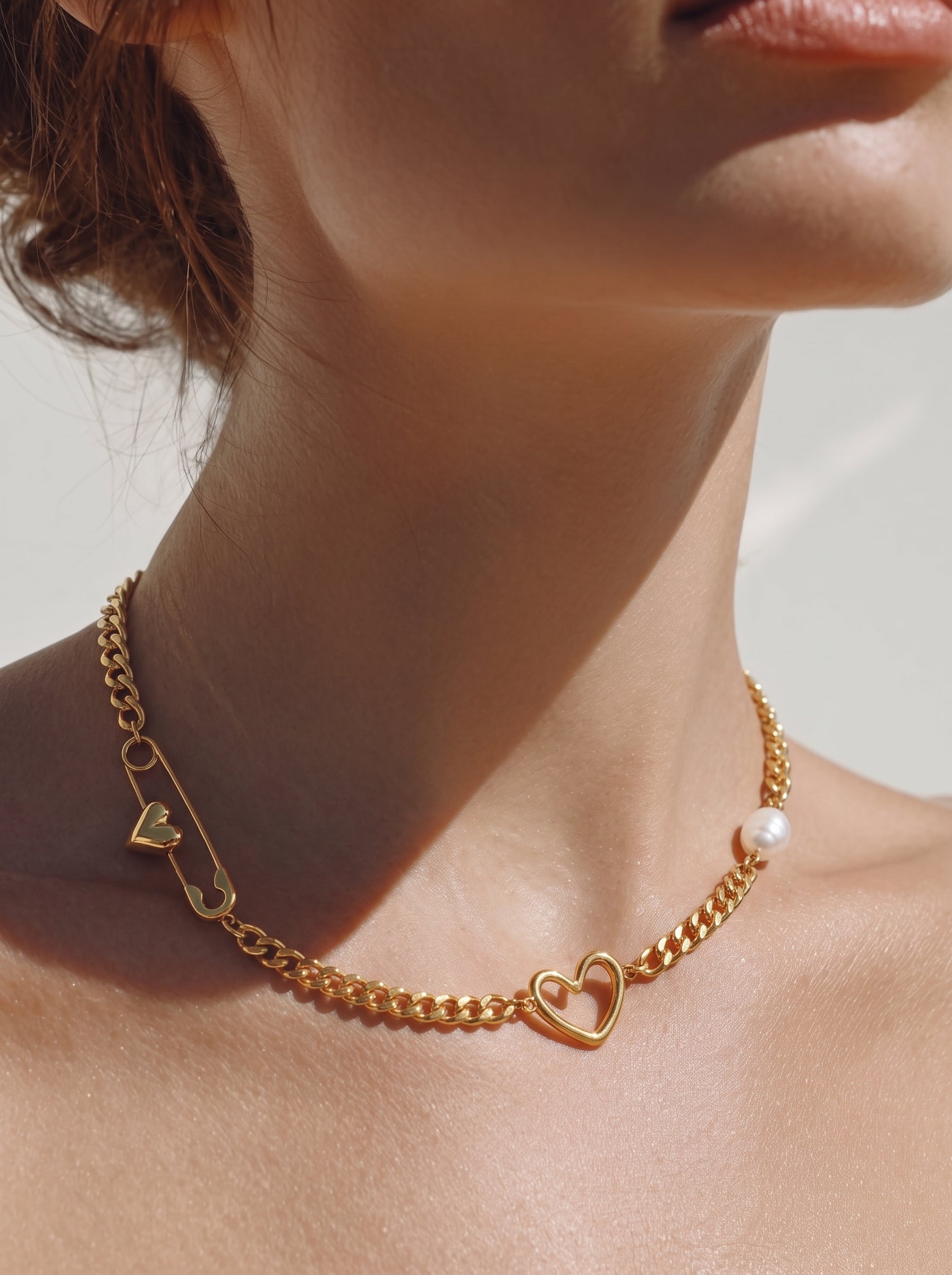 Collier ras-de-cou à chaîne gourmette imposante ton or présentant une breloque épingle de sûreté avec un petit cœur, une breloque cœur ouvert, et une seule perle blanche d'eau douce. Porté par une mannequin. @model