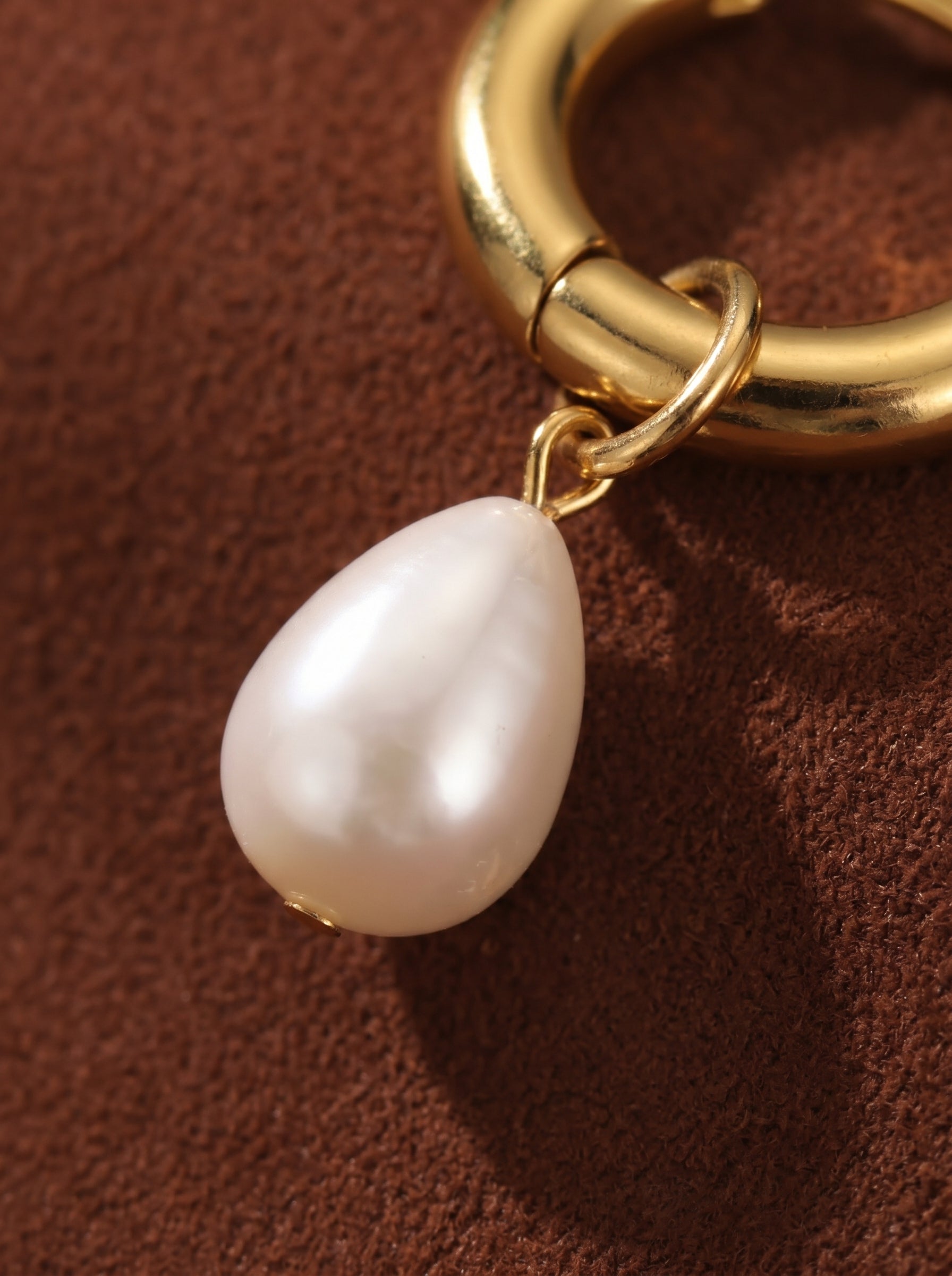 Close-up of a gold hoop earring with a single, large, teardrop-shaped freshwater pearl dangling from it against a brown textured background. @pearl