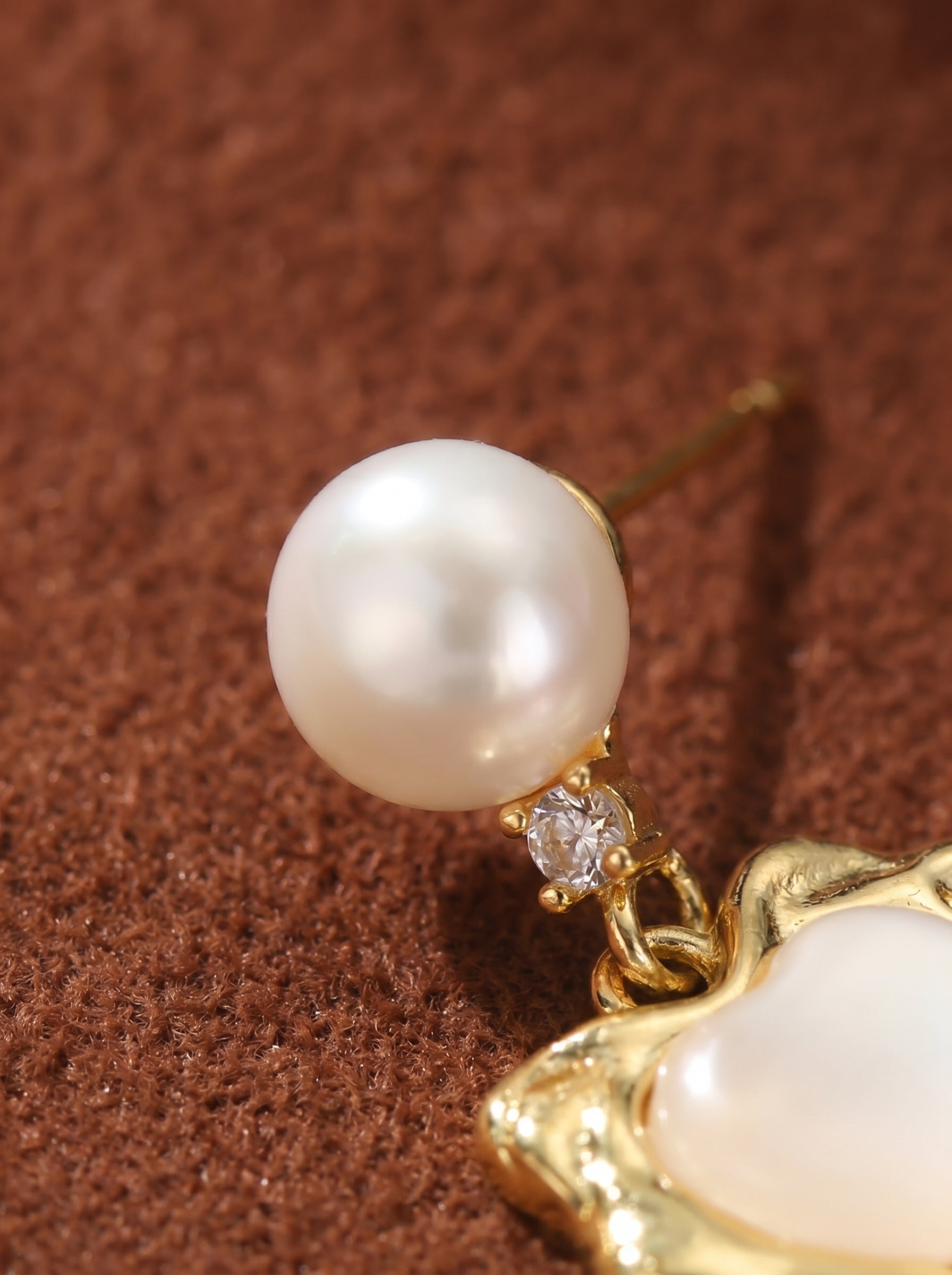 Close-up of a gold-tone stud drop earring featuring a lustrous white freshwater pearl, a small sparkling cubic zirconia accent, and a textured gold heart charm with a white inlay. @pearl