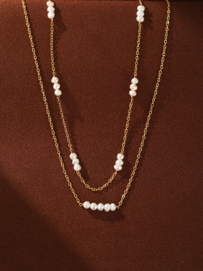 A close-up studio shot of the Stellaris layered necklace, featuring two delicate gold-toned chains adorned with small, luminous white freshwater pearls spaced along their lengths, set against a warm brown background.