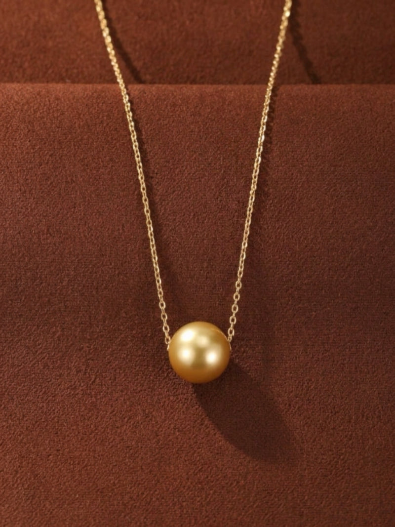 A single, lustrous golden South Sea pearl hangs from a delicate gold chain, presented against a rich brown textured background. The necklace features a minimalist design, highlighting the natural beauty and glow of the pearl.