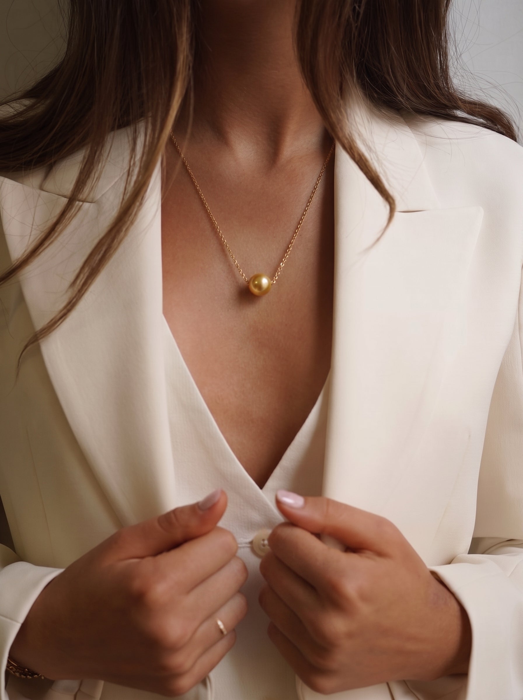 A woman wearing a cream-colored blazer and a white top, adorned with a delicate gold chain necklace featuring a single, lustrous South Sea golden pearl pendant. The image captures a professional and elegant aesthetic, suitable for business attire. @lifestyle