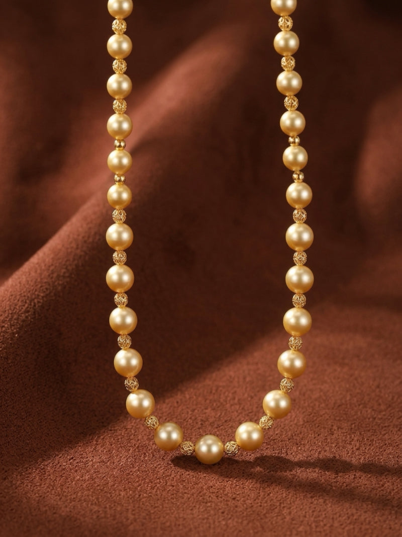 The Sunlit Strand necklace features a strand of lustrous golden freshwater pearls, interspersed with small gold beads, draped elegantly against a rich brown textured background.