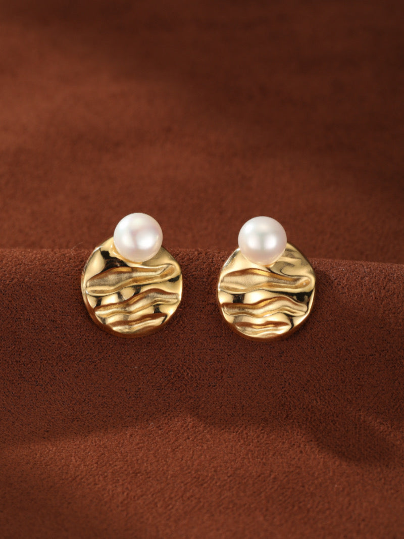 Une paire de boucles d'oreilles puces Surge en perle d'eau douce présentant une perle blanche ronde de 4 mm sertie sur un disque martelé ton or à la surface texturée façon vague, présentée sur fond de tissu marron.