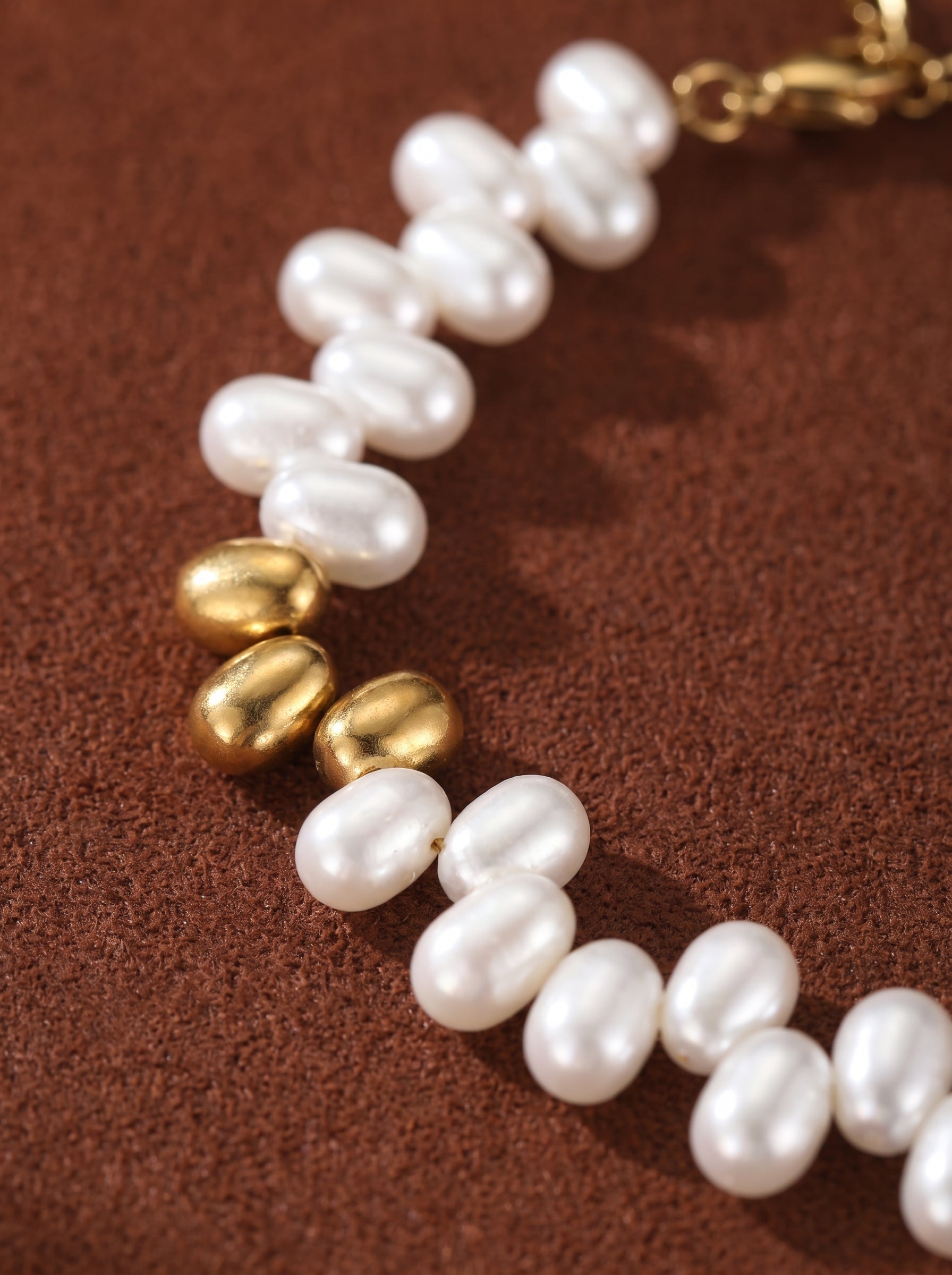 Close-up of a Sylvan bracelet featuring lustrous white freshwater seed pearls and polished gold-toned beads, arranged in a flowing pattern on a textured brown background. @pearl
