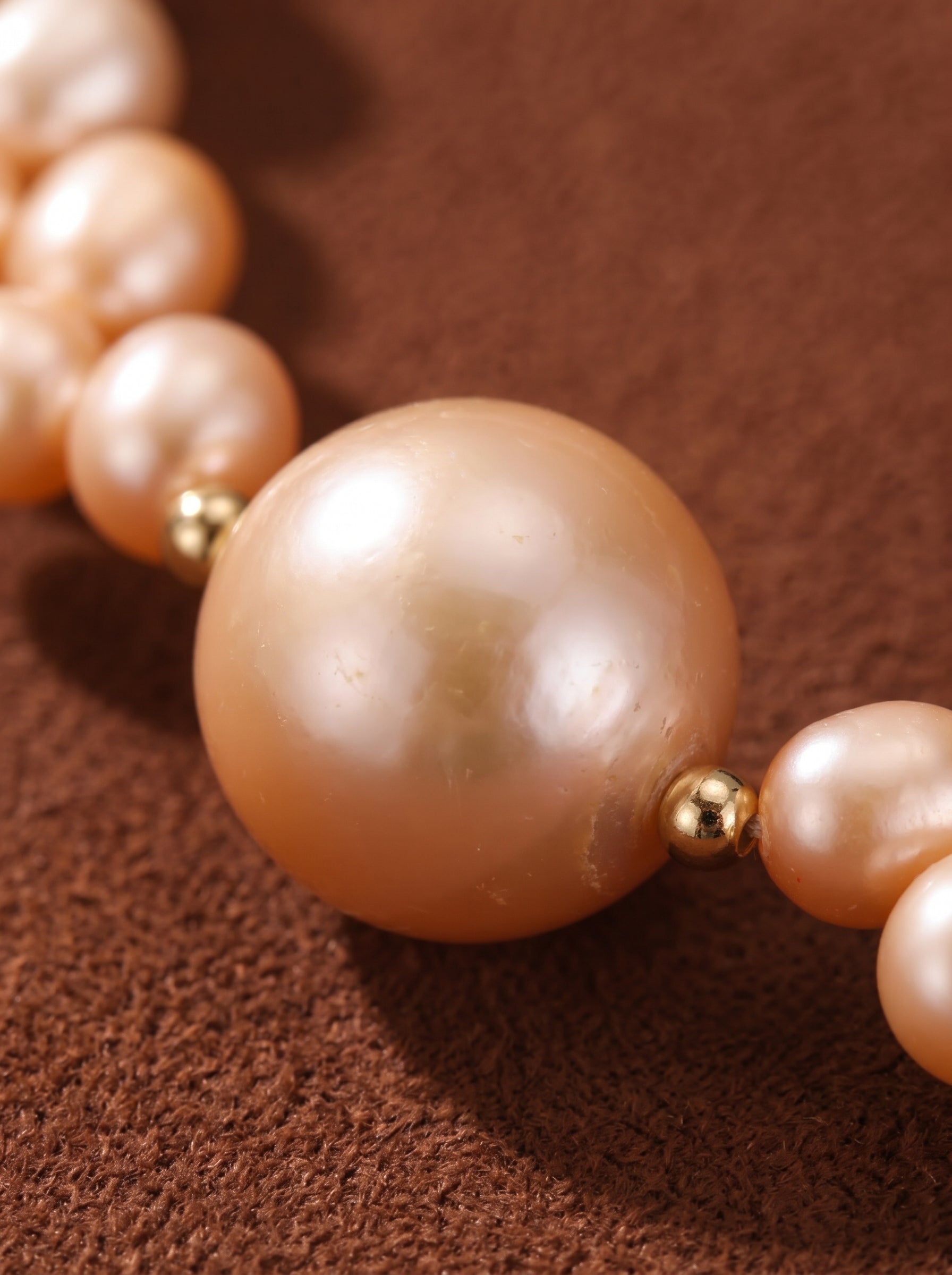 Close-up of a Tendril Freshwater Pearl Beaded Choker Necklace featuring a large, lustrous pink round pearl accented by smaller pink seed pearls and gold beads against a brown textured background.@pearl