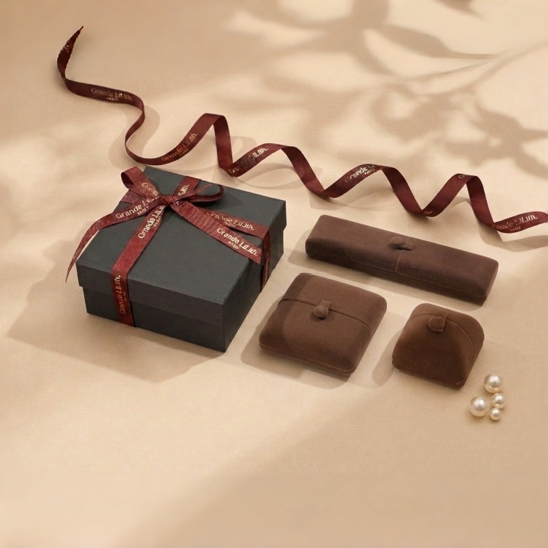 A dark grey gift box tied with a burgundy ribbon featuring the brand name. Beside it are three brown velvet jewelry boxes and a few pearls. @shared