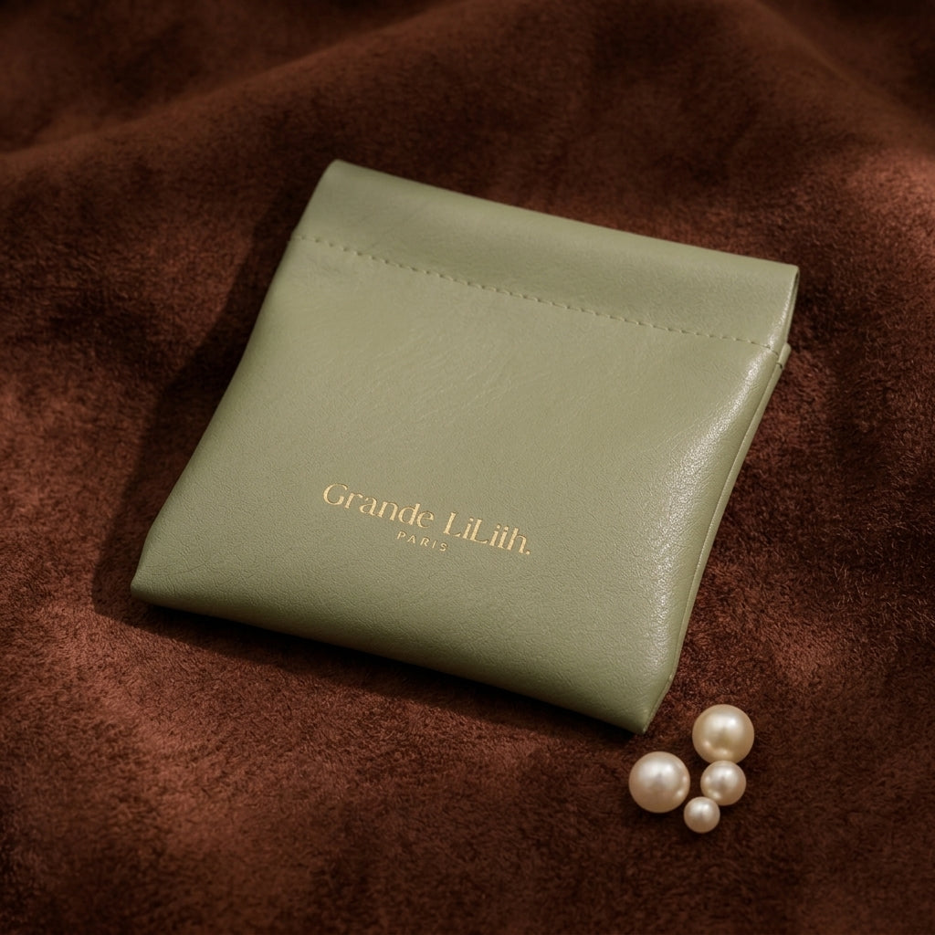 A soft, sage green magnetic pochette with gold embossed "Grande Lilith" logo sits on a brown textured surface next to a few pearls. @shared