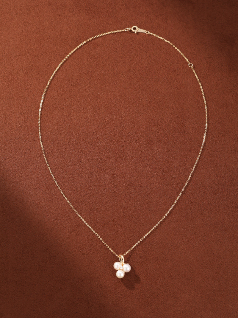 A delicate gold chain necklace featuring a pendant with a cluster of three small, round freshwater pearls on a brown background.