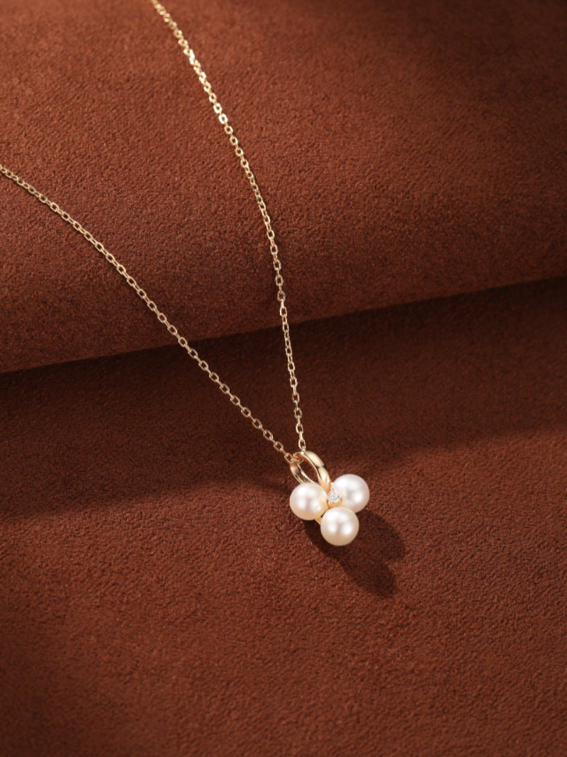 A delicate gold chain necklace featuring a pendant with three luminous 6mm freshwater pearls clustered together, accented by a sparkling stone, set against a rich brown textured background. @pearl