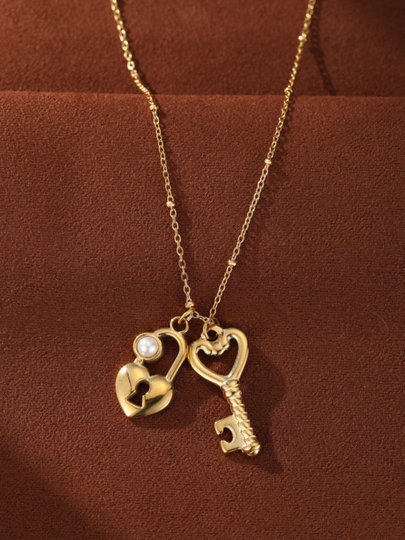 Collier ton or présentant des pendentifs cadenas et clé entrelacés en forme de cœur, avec une petite perle blanche d'eau douce accentuant le cadenas.