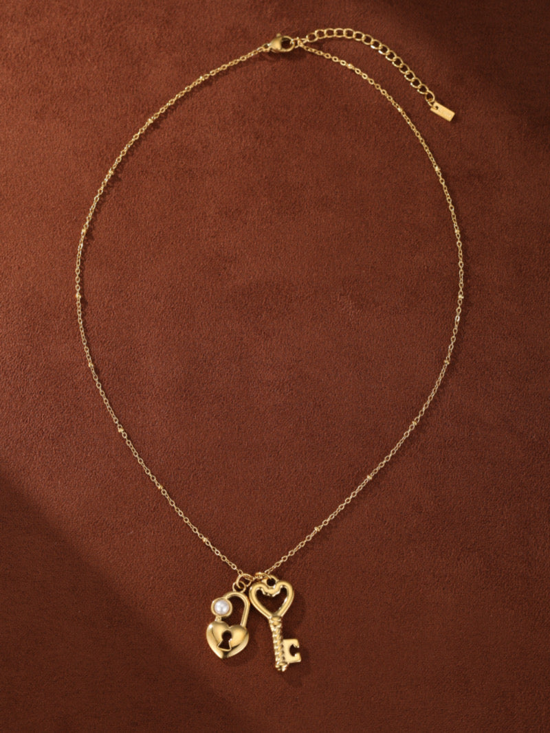 Un collier ton or présentant deux pendentifs en cœur entrelacés : l'un un cadenas orné d'une perle blanche d'eau douce, et l'autre une clé. La délicate chaîne présente de petites perles espacées le long de sa longueur.