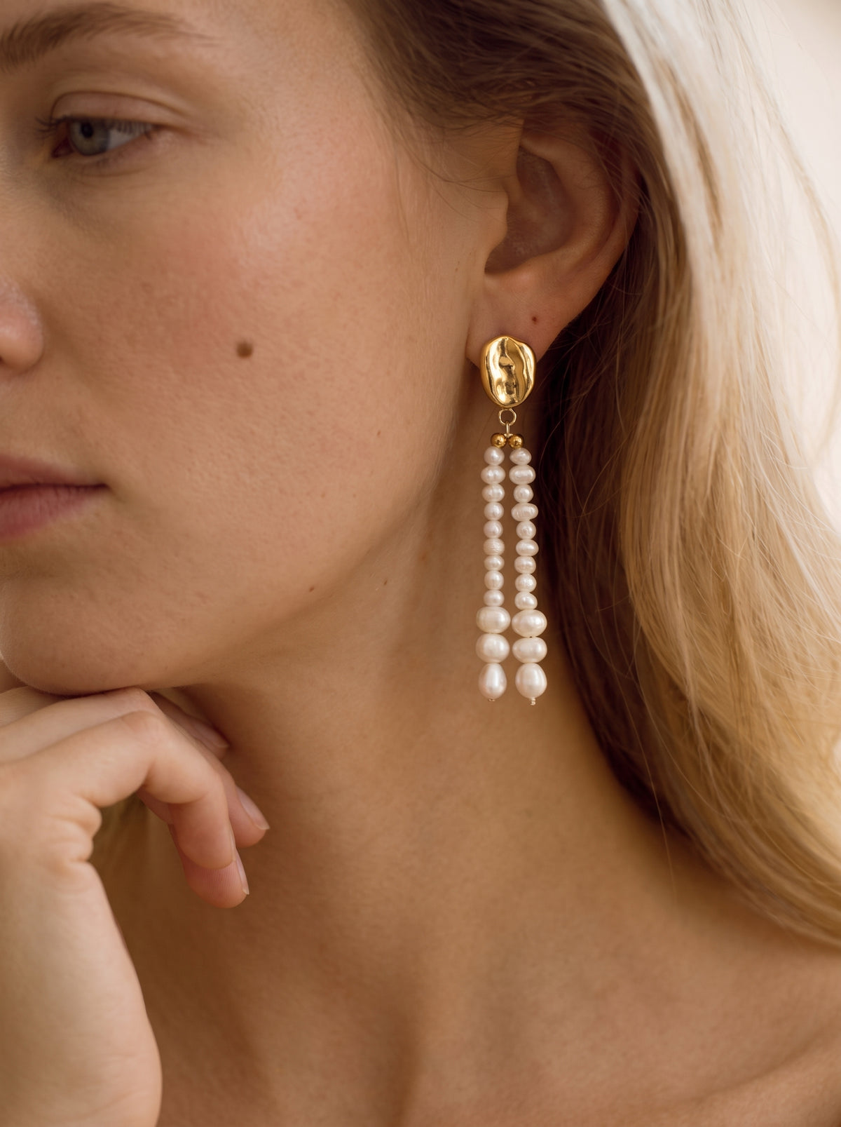 Close-up of a woman wearing Undulate Freshwater Pearl Dangle Earrings with textured gold studs and cascading white pearls of varying sizes. @model