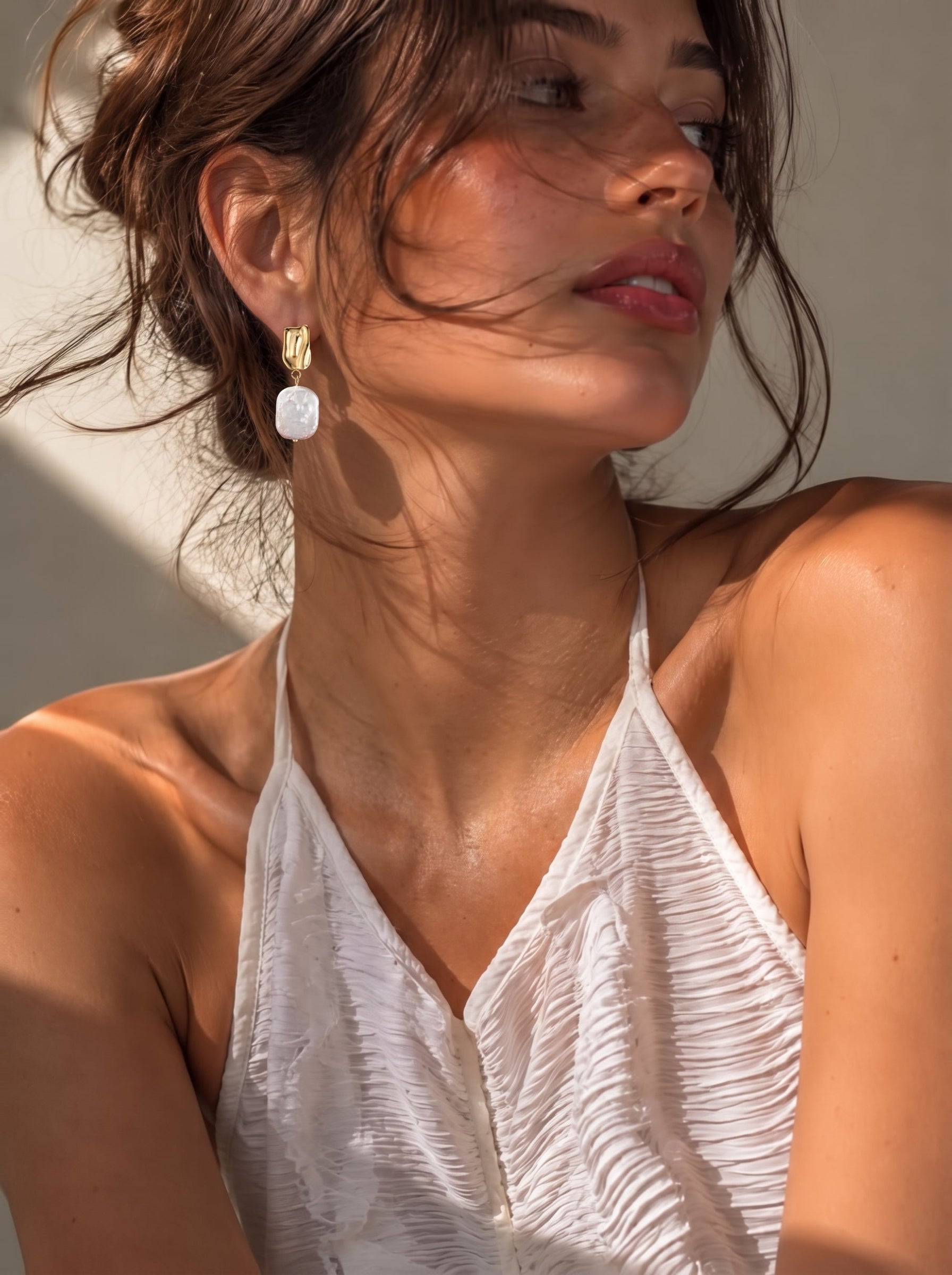 Une femme portant les boucles d'oreilles pendantes Undulate en perle d'eau douce, présentant une puce ton or et une perle baroque pendante, dans un cadre à la lumière naturelle et douce. @lifestyle
