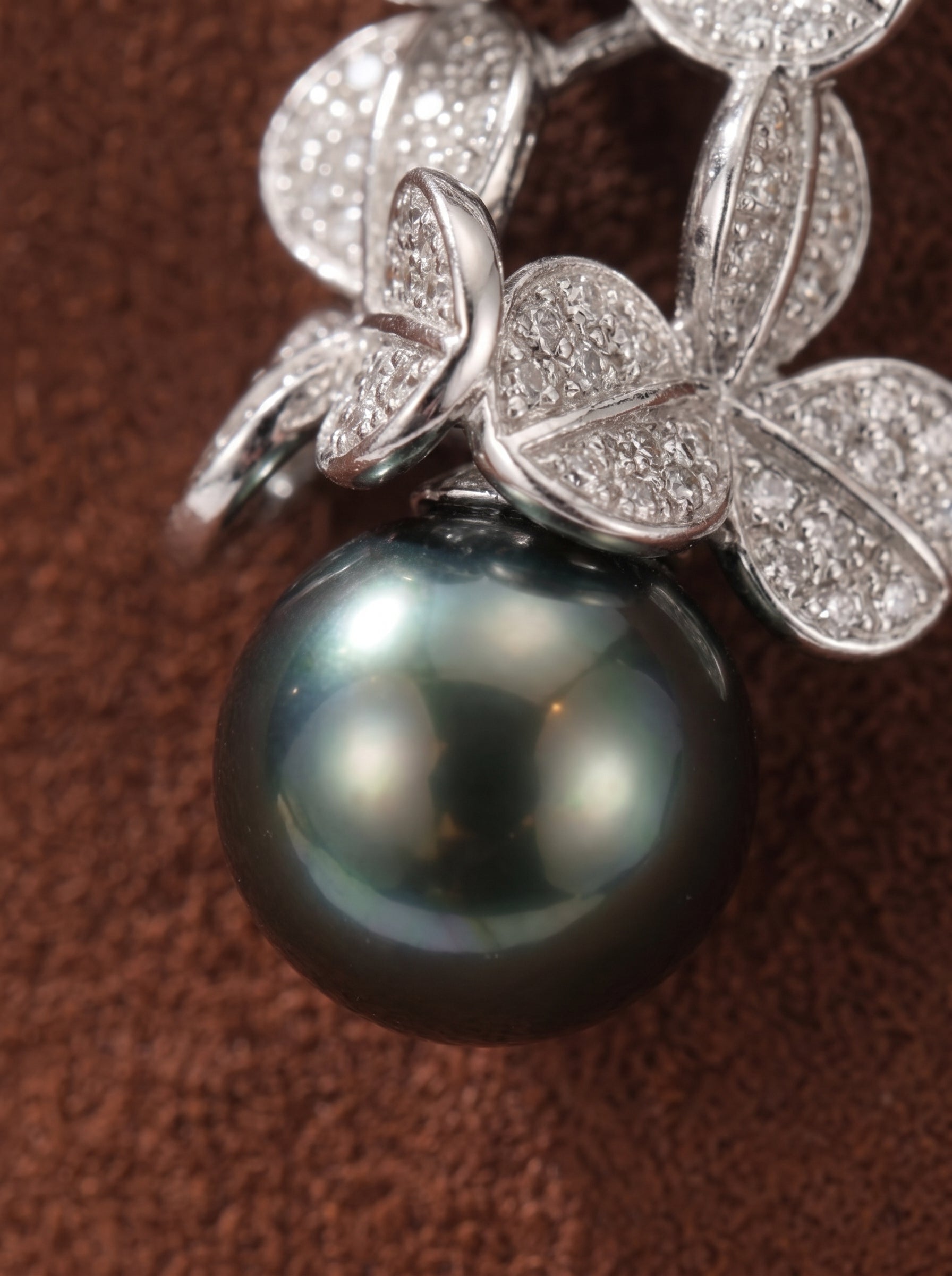Close-up of a pendant necklace featuring a lustrous, round 14mm Tahitian pearl suspended from a cluster of diamond-accented clover-shaped motifs, set against a brown textured background. @pearl