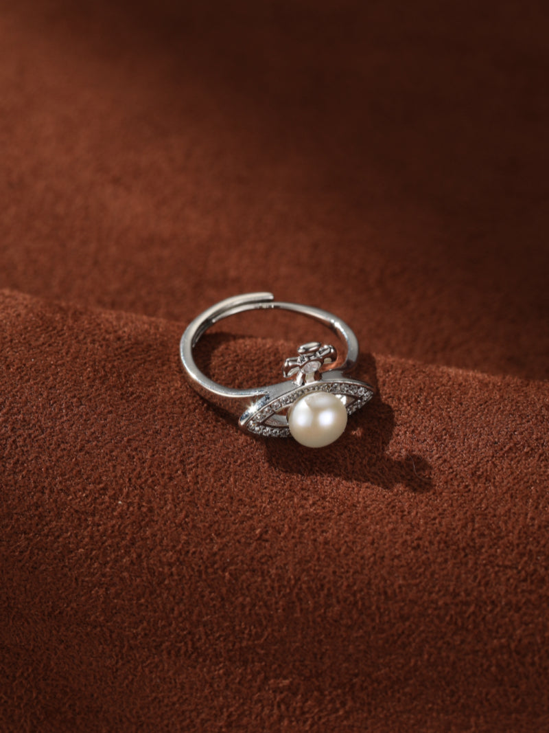 Silver open cuff ring with a central white freshwater pearl, flanked by a cross motif and pave-set crystals on a textured brown surface. @pearl