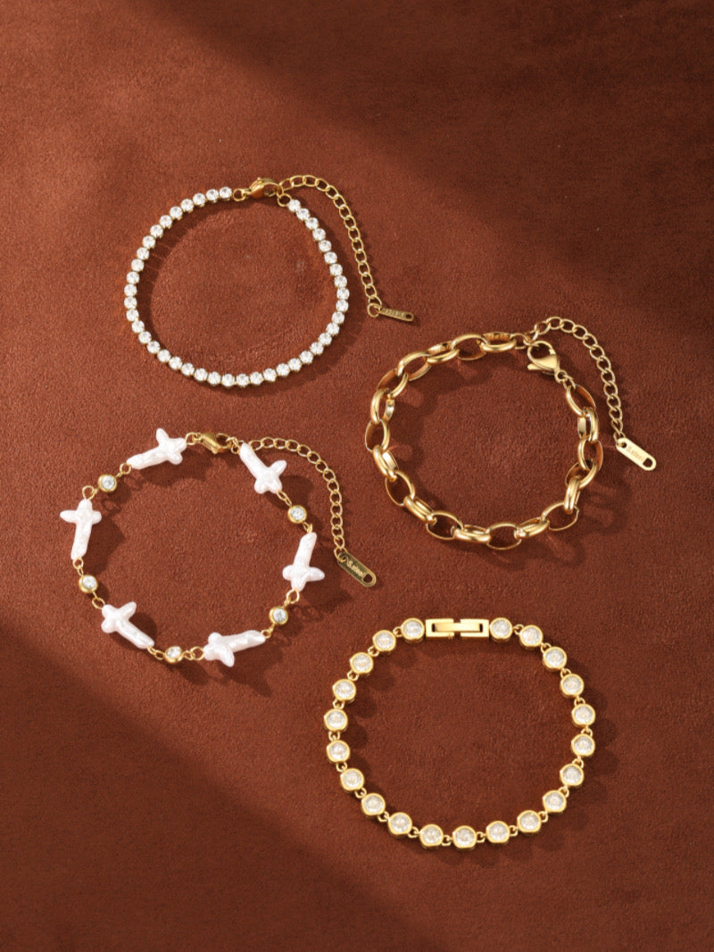 Four gold-toned bracelets displayed on a brown background. One features white cross-shaped freshwater pearls and clear crystals, another is a thick chain, and the other two are delicate chains with clear crystals.