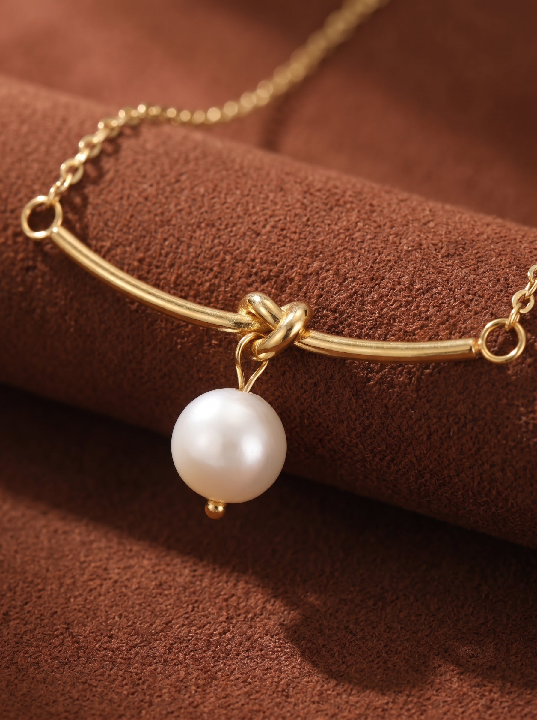 Close-up of a gold chain necklace featuring a single white freshwater pearl suspended from a curved bar, secured by a knot detail. Verve collection.@pearl