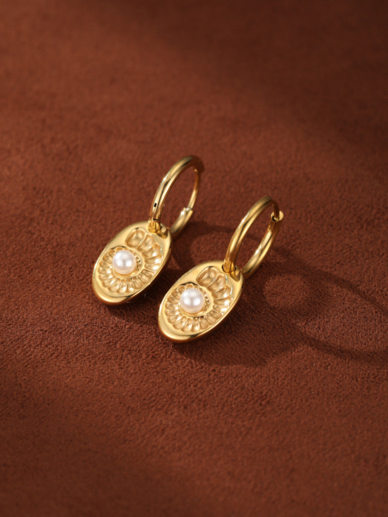 Gold hoop drop earrings featuring an oval charm with a white freshwater button pearl at its center, embossed with a delicate spiral pattern, displayed on a textured brown surface.