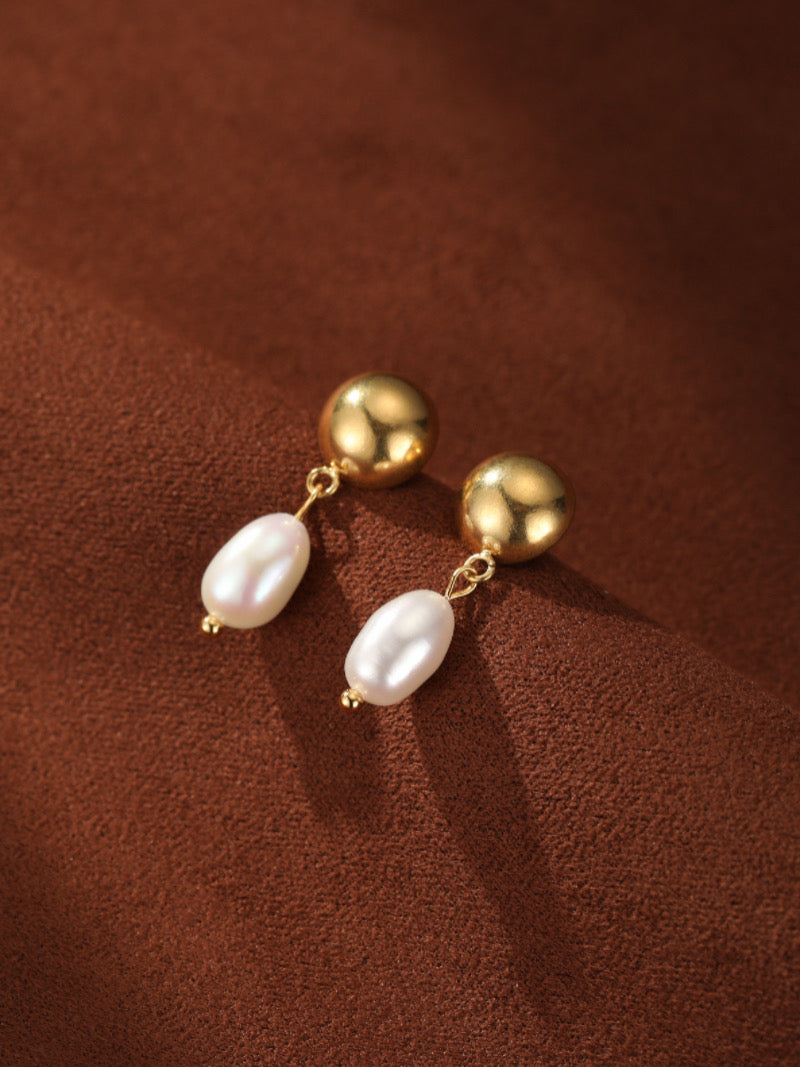 Boucles d'oreilles goutte en perle d'eau douce Vesper avec une sphère dorée polie et une lustrée perle baroque blanche suspendue en dessous, présentées contre un fond texturé marron.