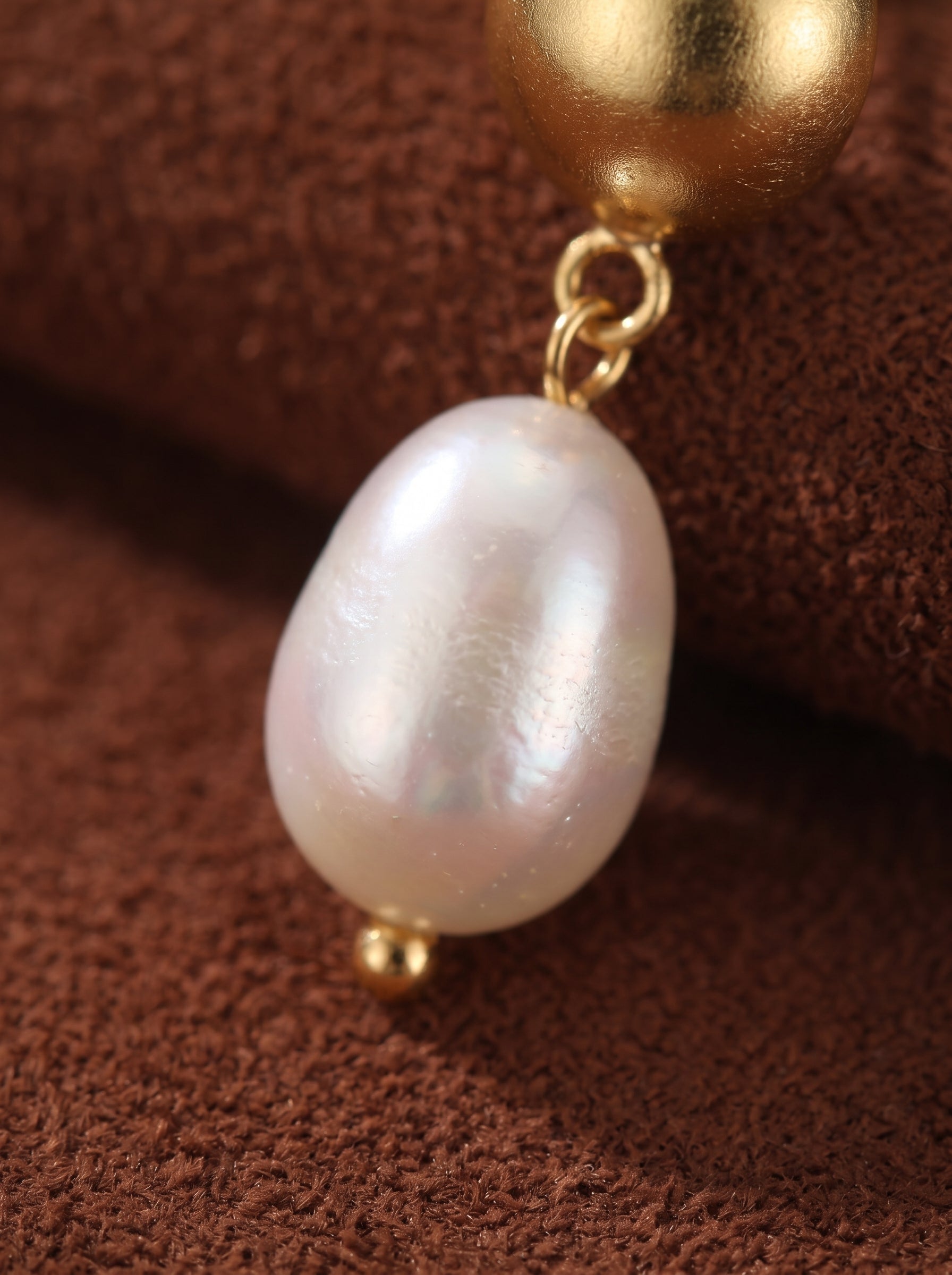 Photo en gros plan des boucles d'oreilles Vesper, révélant une lustrée perle blanche d'eau douce en forme de goutte suspendue à une sphère dorée texturée. @pearl