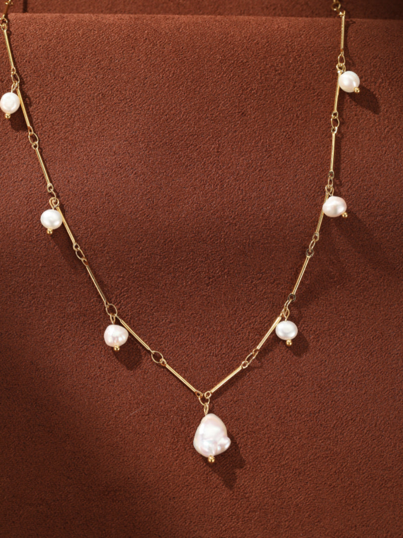 A delicate gold-tone chain necklace featuring multiple small freshwater pearls and a large baroque pearl pendant, displayed on a textured brown background. @pearl