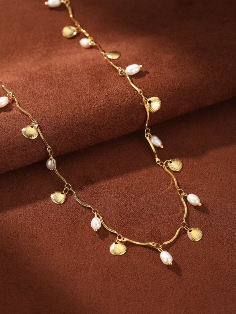 A gold-tone necklace with a wavy chain, alternating small white freshwater pearls and tiny seashell charms, displayed on a textured brown background. @pearl