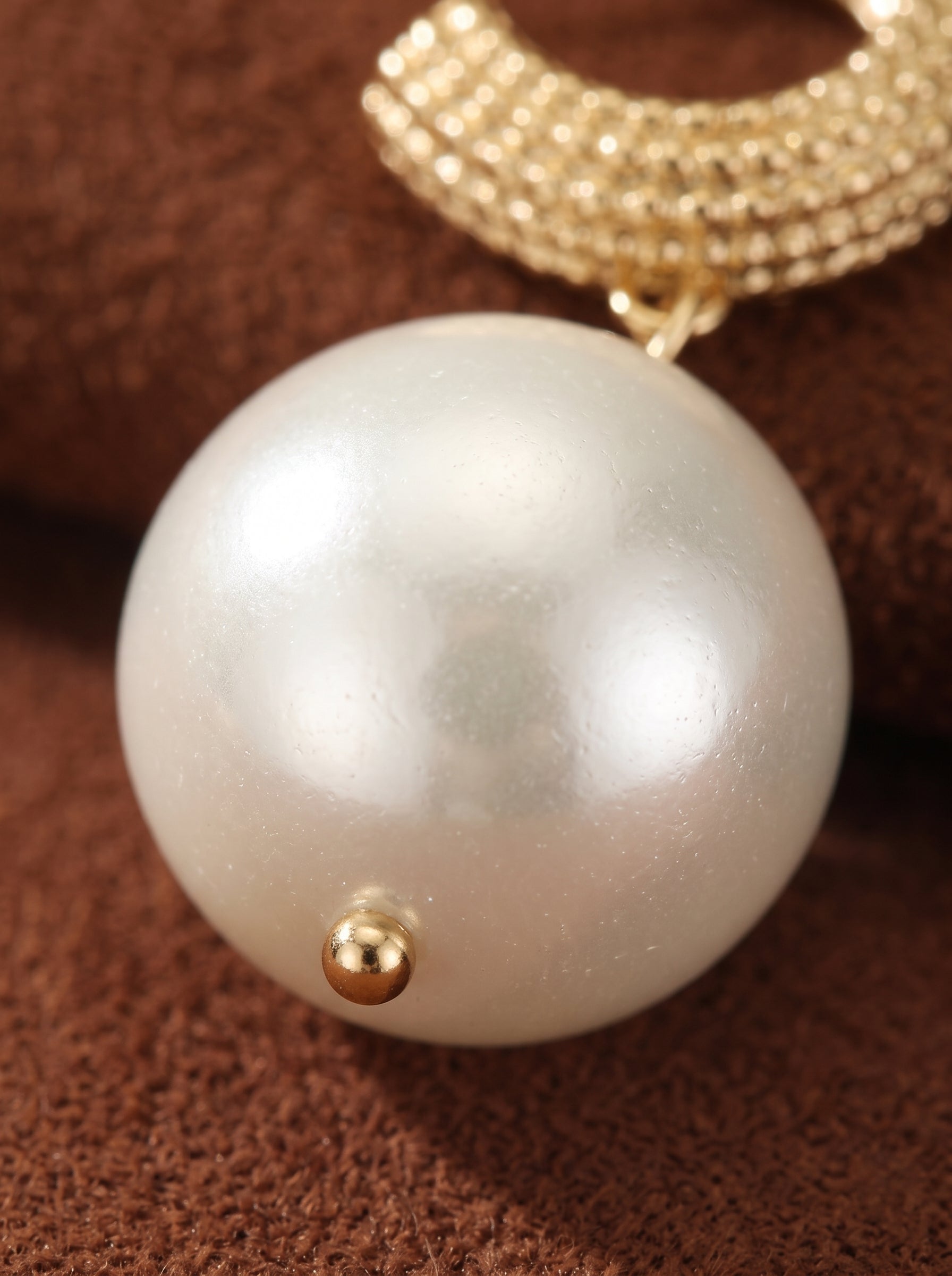 Close-up of a large, round white imitation pearl drop earring with a textured gold-toned hoop detail and a small gold bead accent, on a brown textured background. @pearl