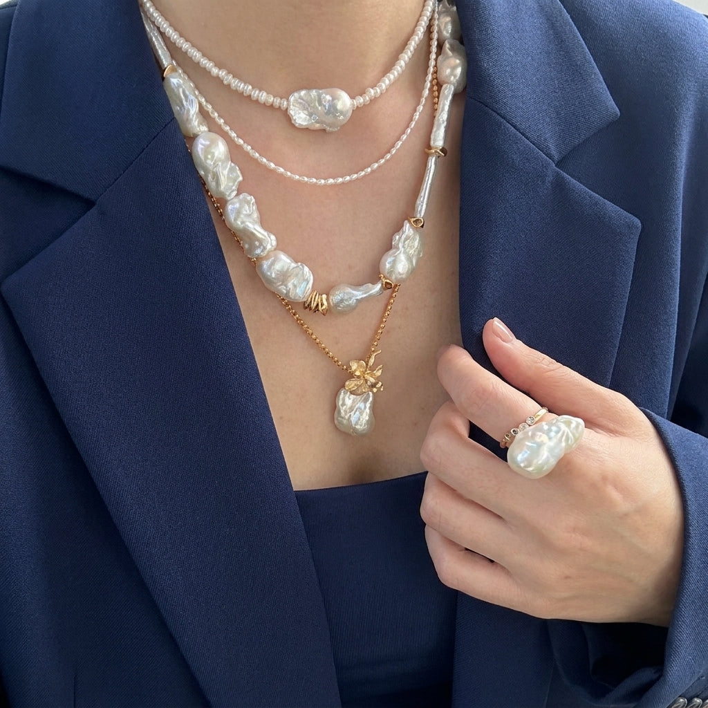 A modern professional woman wearing a layered stack of large, irregular white baroque pearl necklaces, a baroque pearl pendant, and a matching oversized baroque pearl statement ring, styled with a navy blue blazer.