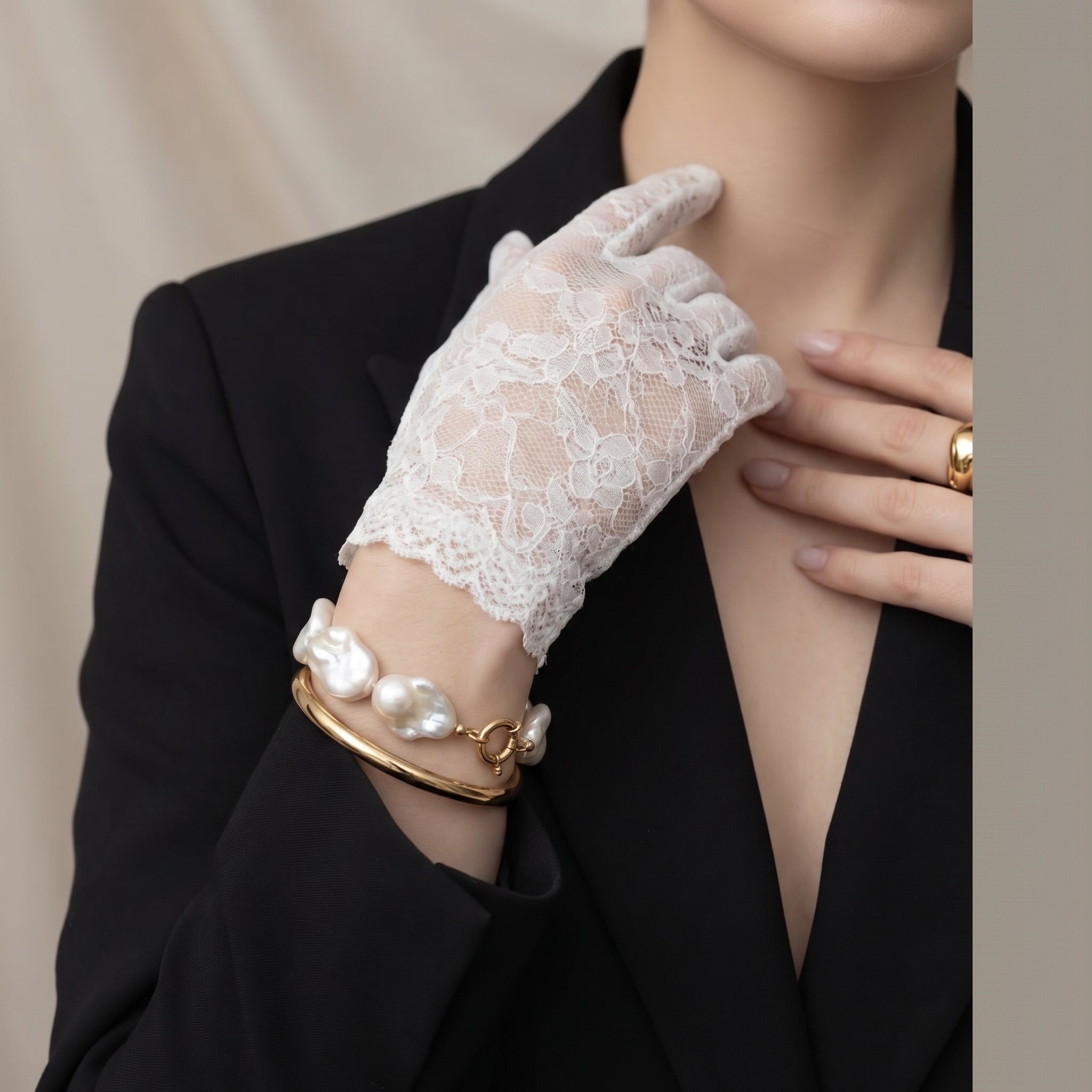 Close-up of a modern woman wearing a black blazer and white lace glove, elegantly styling a Grande Lilith baroque freshwater bracelet stacked with a gold bangle.