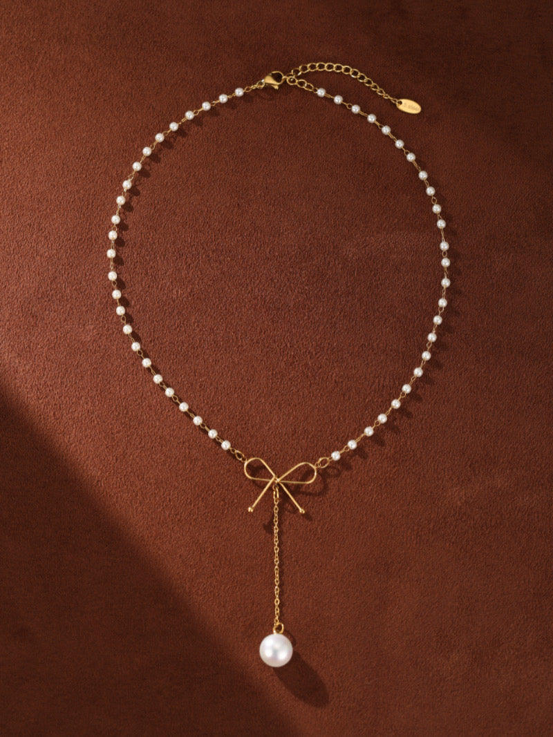 Gold-tone necklace with a delicate chain of small white imitation pearls, featuring a small gold bow pendant with a single larger imitation pearl drop.