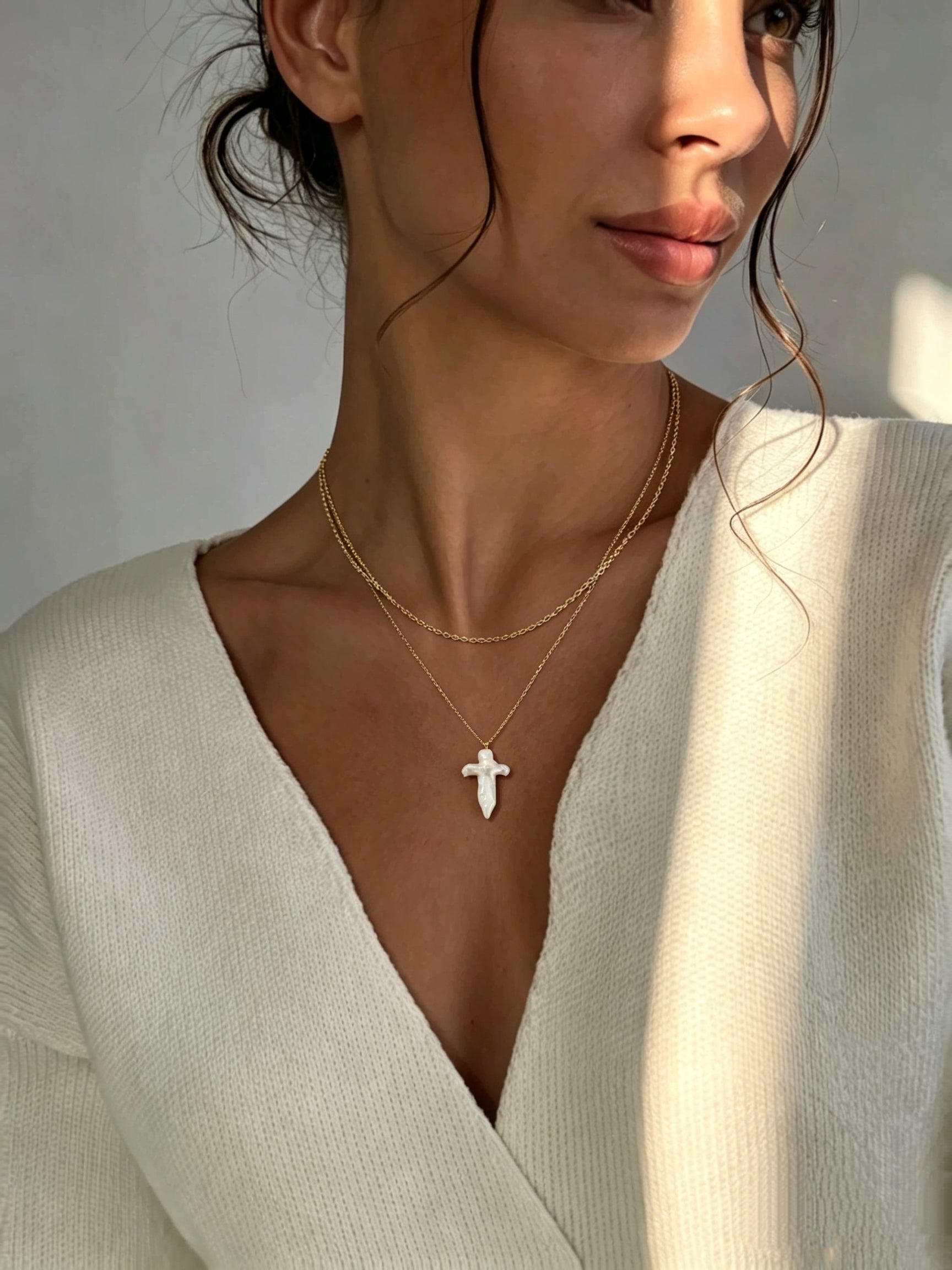 A woman wearing a white sweater and the Aegis layered necklace featuring a large, white baroque freshwater pearl cross pendant on a gold-toned chain. @model