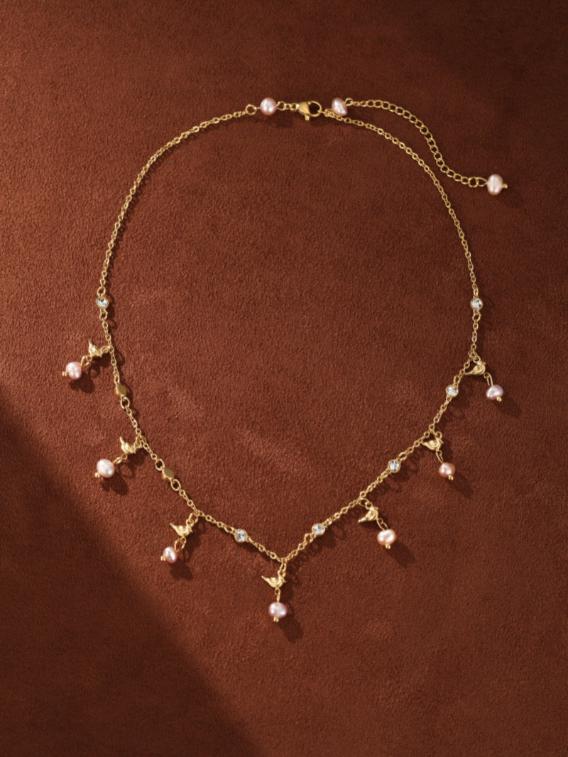 Un délicat collier flottant ton or présentant une chaîne avec de multiples petites perles roses d'eau douce, des motifs d'oiseau, et des accents en cristal transparent.