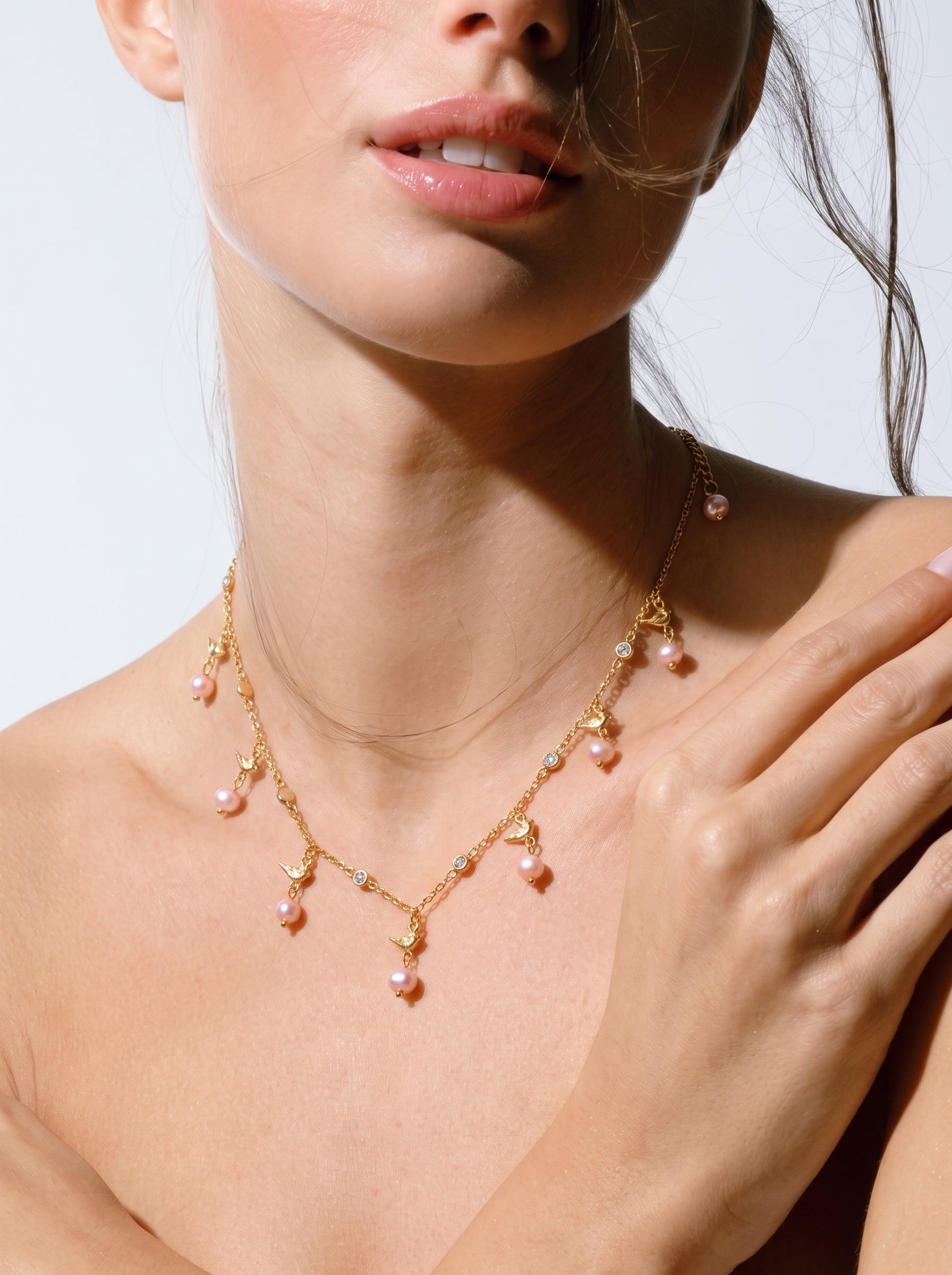 Un délicat collier à chaîne ton or présentant de multiples petites perles roses d'eau douce, de minuscules breloques oiseau, et des accents en cristal transparent, porté par une mannequin. @model
