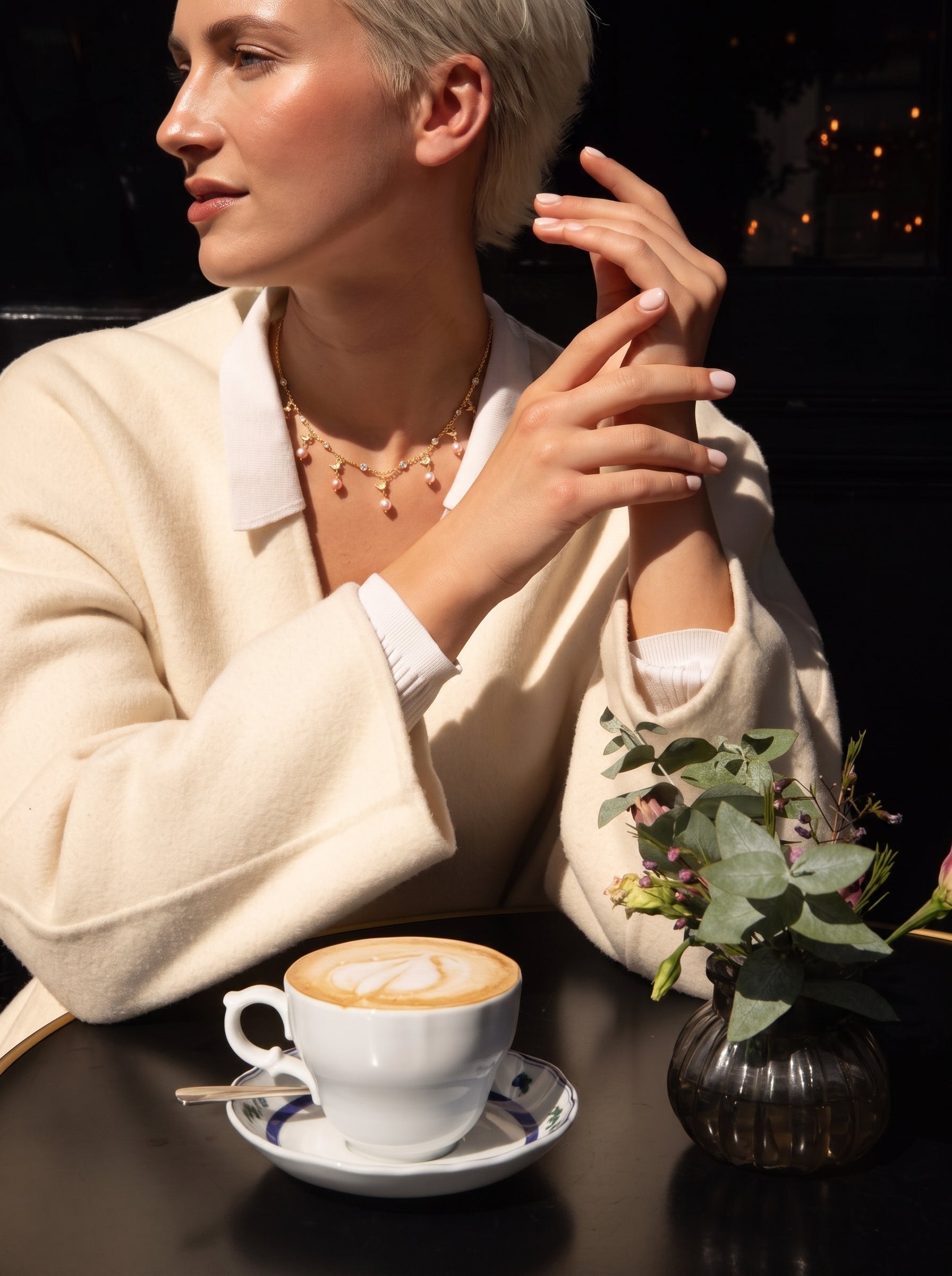 Une femme portant le collier flottant oiseau Aerie en perle d'eau douce tout en savourant un café dans un café. Le collier présente une délicate chaîne ton or avec de petites perles d'eau douce et des motifs d'oiseau. @lifestyle