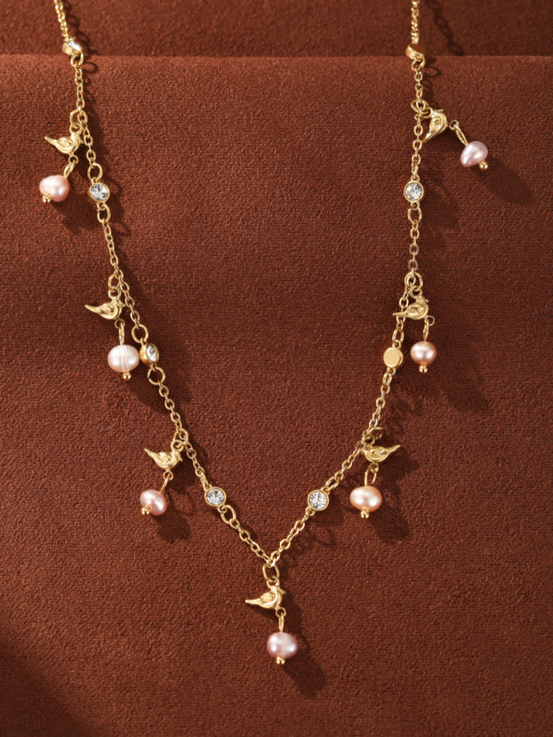 Un collier à chaîne ton or avec de multiples petites perles roses d'eau douce, de délicates breloques oiseau, et des accents en cristal transparent. Le collier a un design flottant et est présenté sur fond marron.