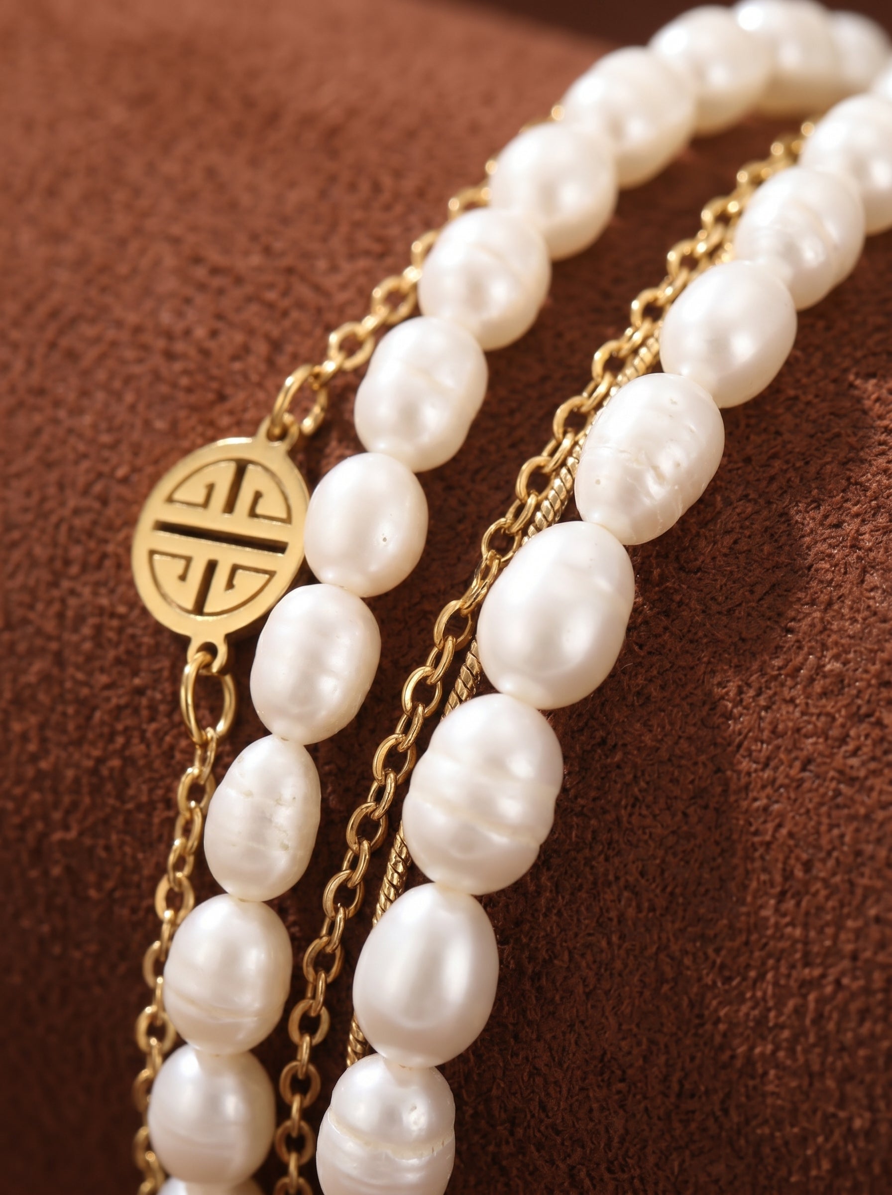 Close-up of a layered bracelet featuring cream freshwater seed pearls and gold-plated stainless steel chains, with a circular gold charm displaying a longevity motif.@pearl