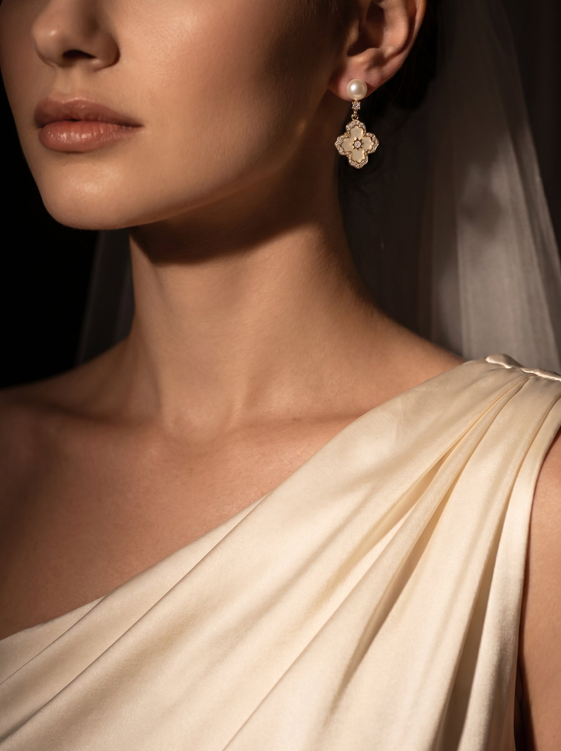 Close-up of a woman wearing Aethel Freshwater Pearl Cross Drop Stud Earrings, featuring a white button pearl above a gold-tone clover-like motif with sparkling stones. @model