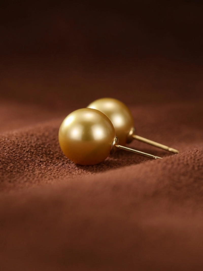 A pair of lustrous golden freshwater pearl stud earrings with gold posts, displayed on a textured brown surface.