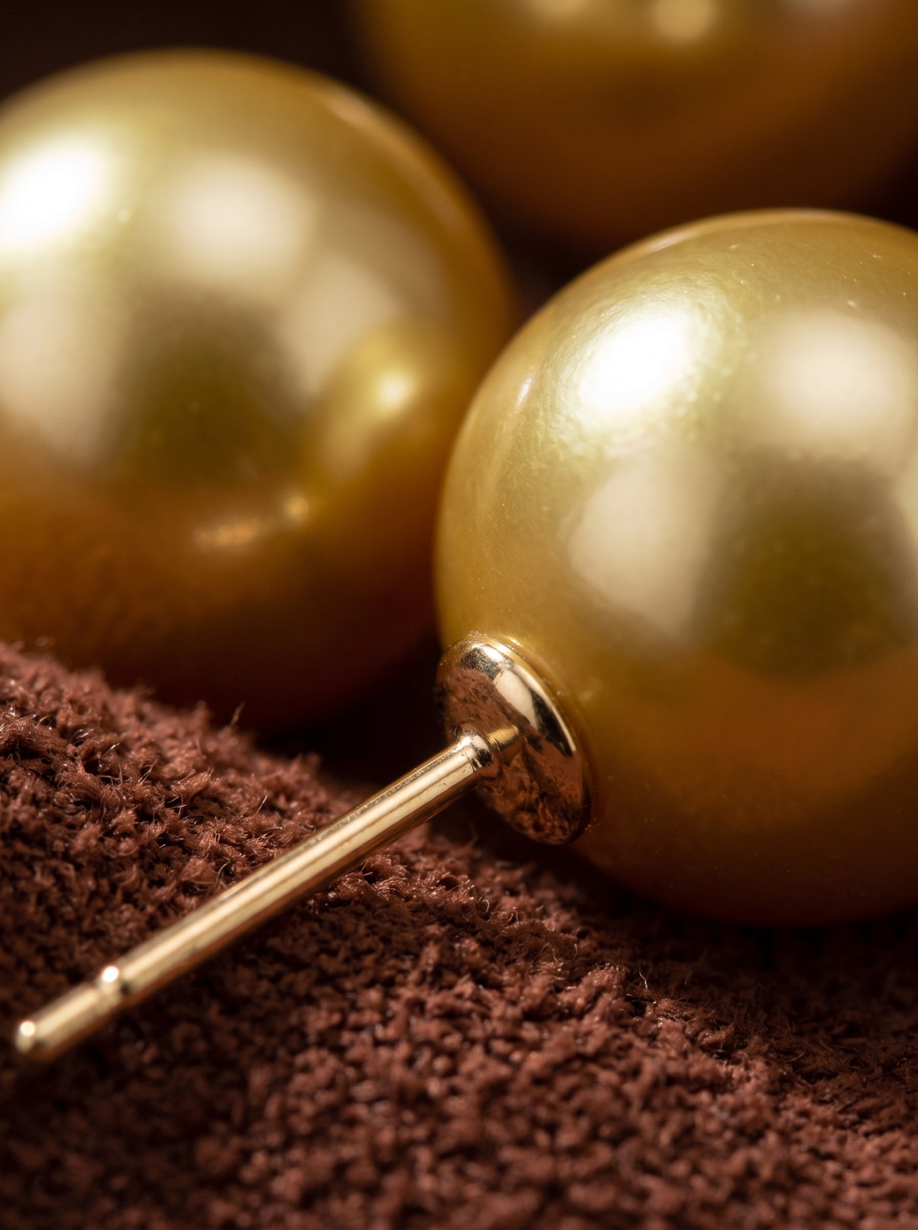 Close-up of two lustrous golden freshwater pearl stud earrings with gold posts, resting on a textured brown surface. @pearl