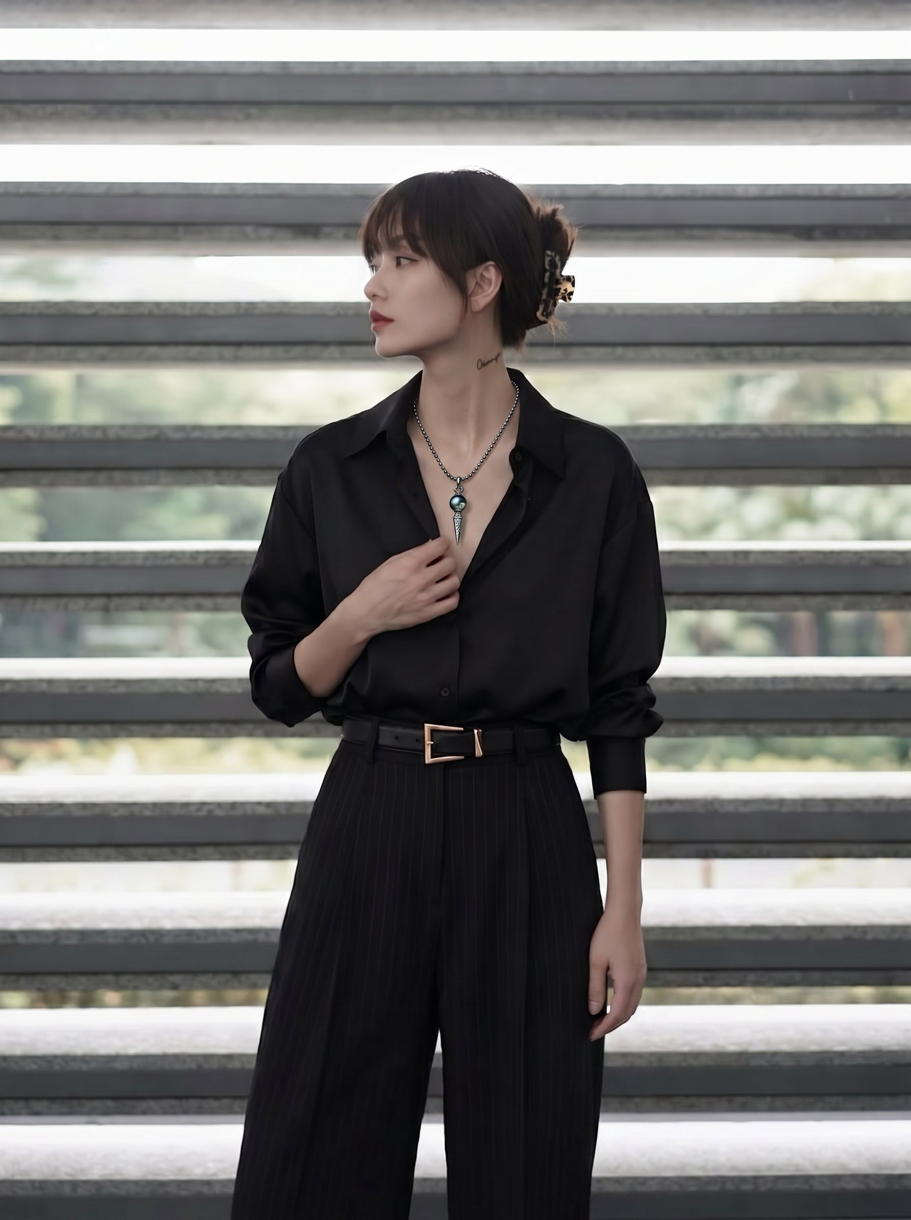 A woman models the Amulet - Tahitian Pearl Phurba Large Drop Necklace, featuring a black pearl and a phurba pendant, against a blurred background of horizontal steps. @model #lifestyle