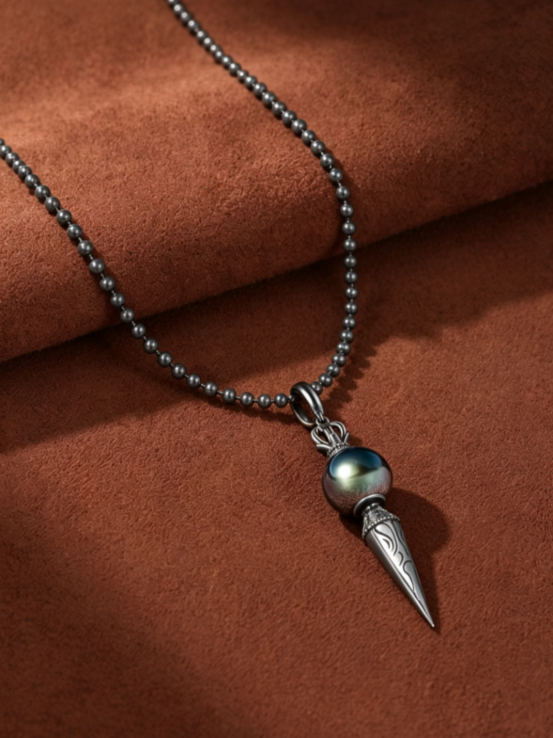 Amulet necklace featuring a large, round Tahitian pearl with iridescent green and blue overtones, suspended above a detailed silver Phurba pendant with a spiked, patterned design. The necklace is on a dark ball chain and presented against a textured brown background. @pearl