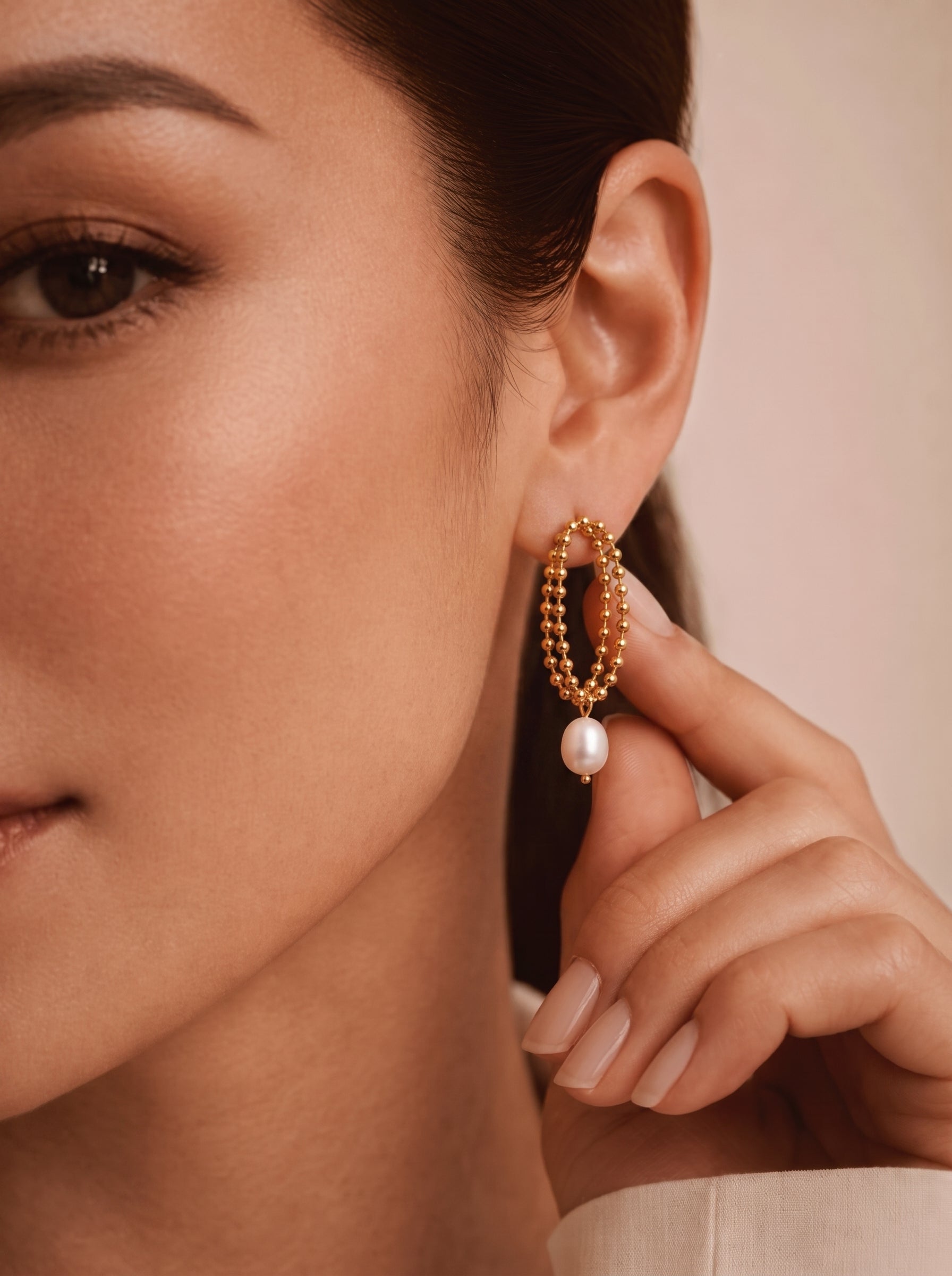 A woman's ear adorned with gold-toned Aperture Freshwater Pearl Drop Earrings, featuring a double-strand beaded oval design and a single lustrous pearl drop. @model